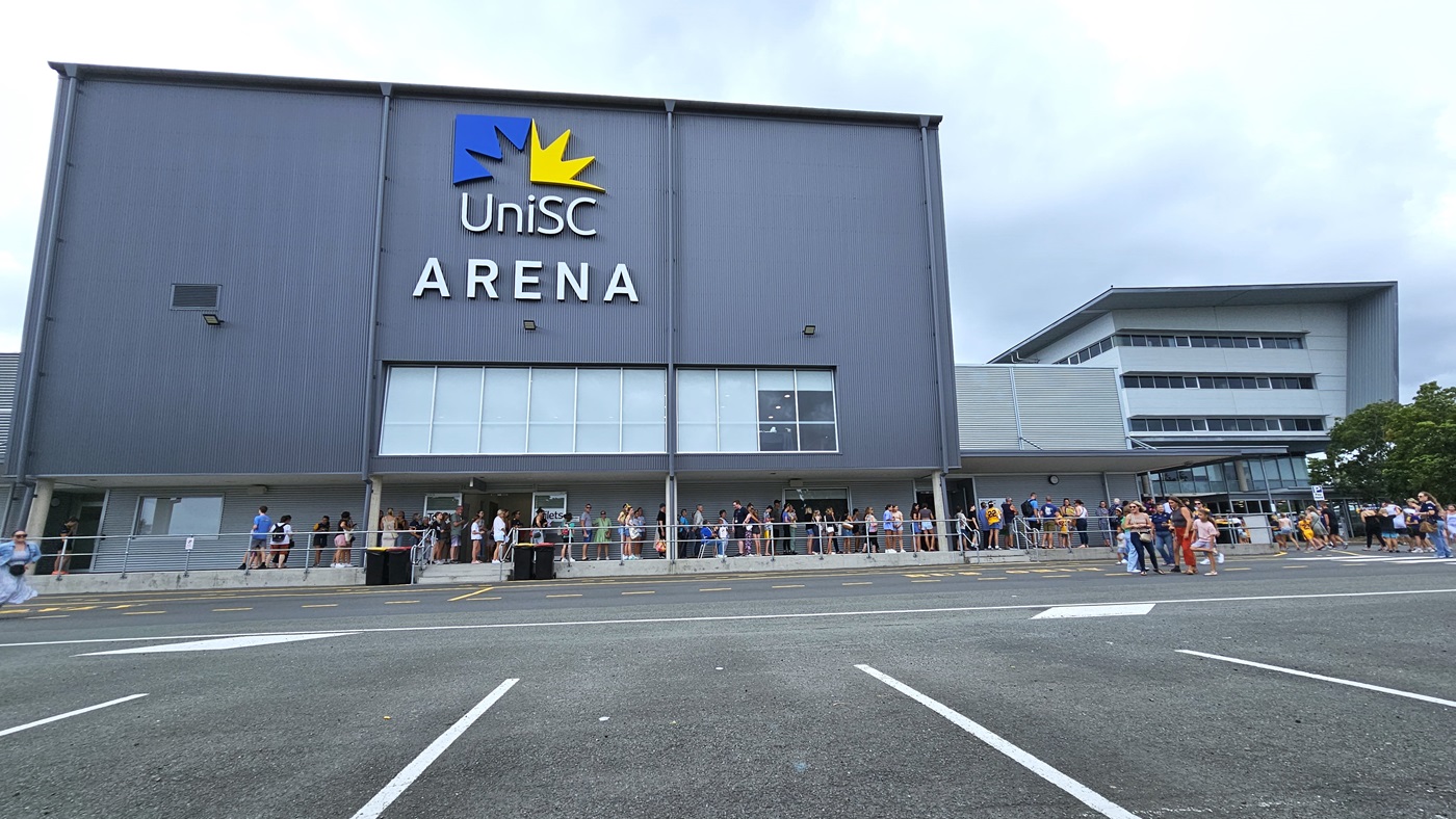 A large grey building with a "UniSC Arena" sign and a yellow star logo. A long queue of people is lined up along the front of the building, waiting to enter. In the middle, the entrance to the arena has glass doors and windows, with more people gathered around. The right side features an extension of the building with additional windows and a covered walkway leading to another entrance. The foreground shows an asphalt car park with white lines marking parking spaces.