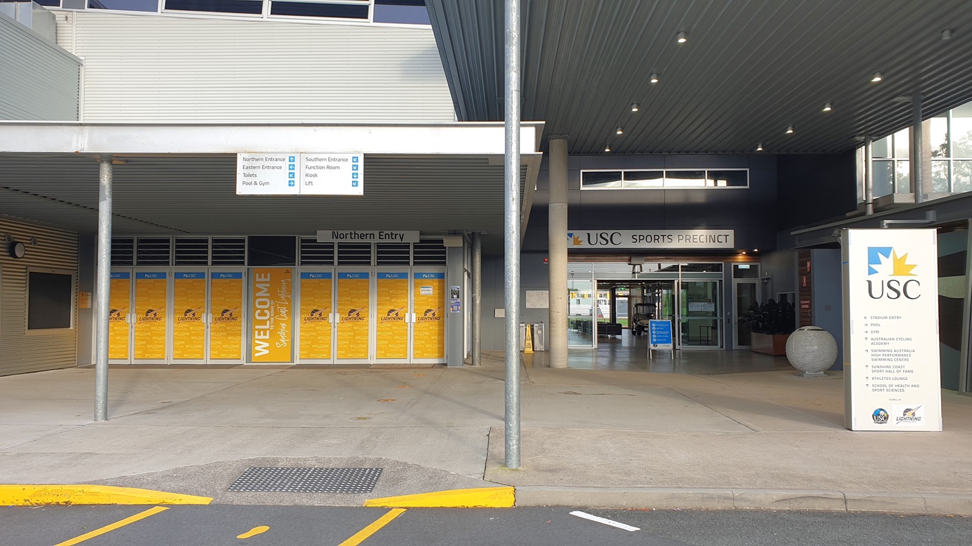 On the left, there is a grey wall with yellow posters and a "Northern Entry" sign above. The middle section has a covered entrance with a "Welcome" sign and glass doors leading inside. The right side features another entrance with a "USC Sports Precinct" sign above the doors. The foreground shows a concrete pavement with a yellow kerb and a metal grate. The ceiling is covered with grey panels and has recessed lighting. The background includes more of the building's facade and windows.