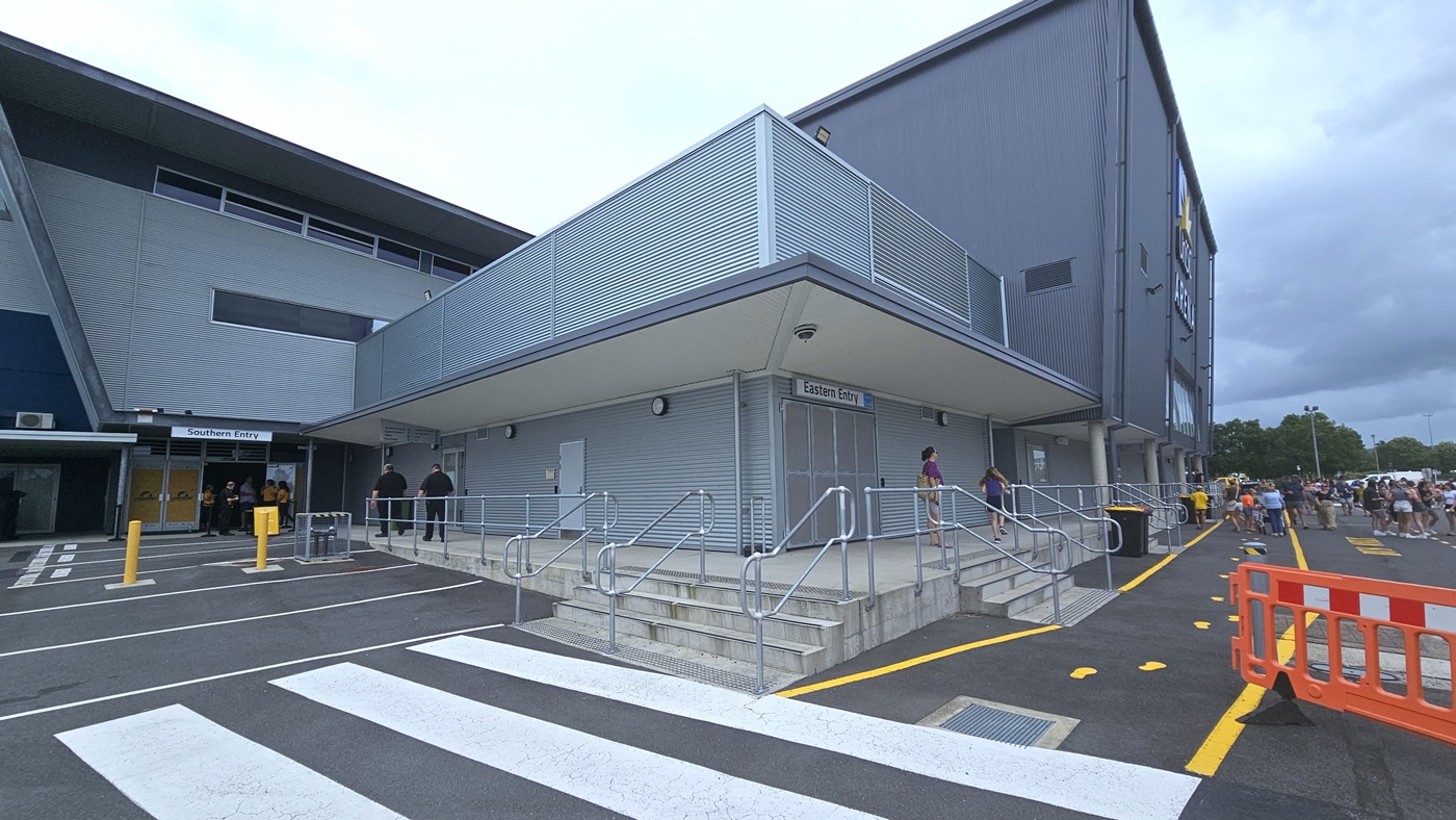 On the left, there is a grey building with a "Northern Entry" sign above a glass door. The middle section features a covered entrance with a ramp and stairs with handrails, leading to double doors with a "Sports Centre" sign. The right side shows a continuation of the building with additional windows and a crowd of people. The foreground includes a zebra crossing on an asphalt surface, yellow kerbs, and a metal railing along the ramp. Truncated domes are installed before the stairs.