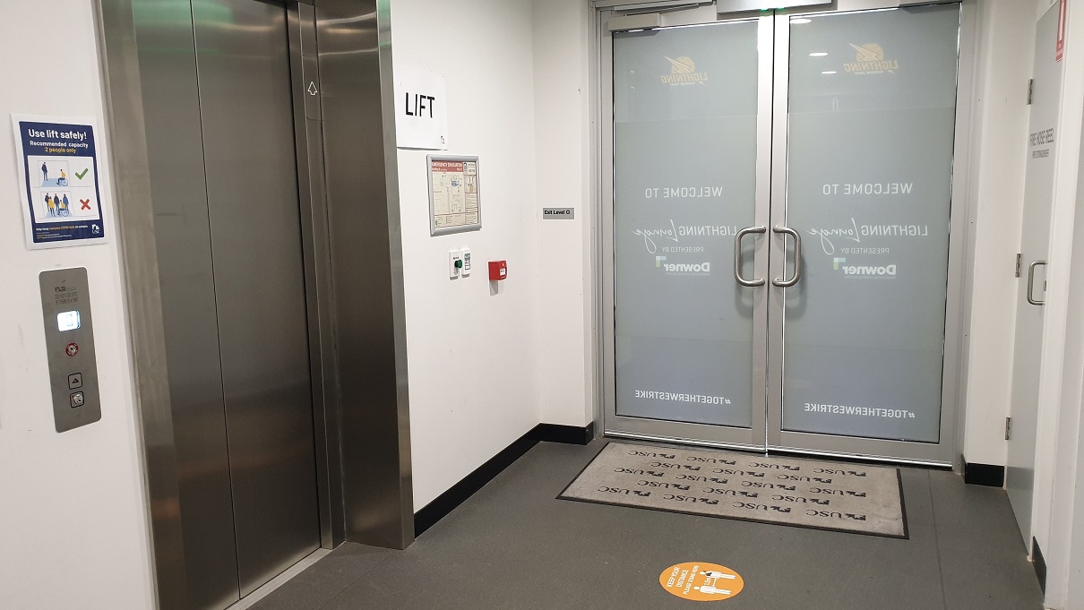 On the left, a lift features stainless steel doors and a control panel, with a "LIFT" sign above. Beside it, a fire alarm and an emergency evacuation plan are on the wall. The middle section presents glass doors with frosted panels marked "WELCOME TO LIGHTNING LOCKER ROOM" and "DOWNER," complete with metal handles and a mat stating "TOGETHER WE STRIKE." On the right, a white wall includes a door to another room. The area is carpeted in grey.