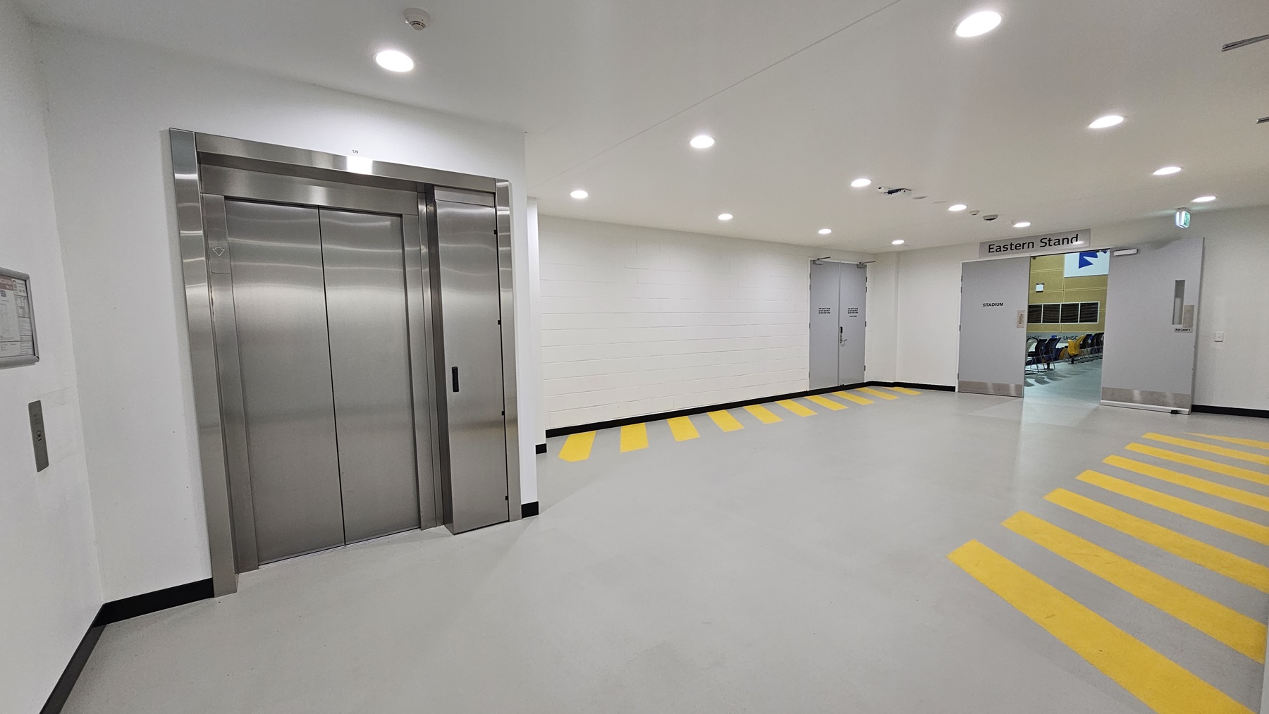 On the left, there is a stainless steel lift with closed doors and a control panel on the wall beside it. The middle section features a white wall with a sign above a door that reads "Eastern Stand." The right side has a corridor leading to another area, with yellow and black striped markings on the floor indicating a pedestrian path. The floor is grey and smooth, and the ceiling has recessed lighting with several lights turned on.