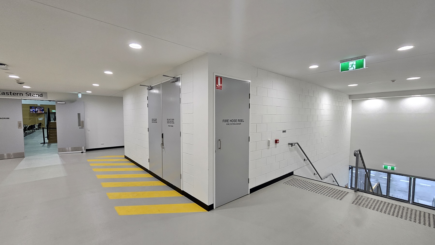 On the left, there is a white wall with a sign reading "Eastern Stand" above a doorway leading to another area. The middle section features two grey doors with "Fire Hose Reel" written on them, and a fire extinguisher mounted on the wall. The right side has a staircase with metal railings and truncated domes descending to a lower level, and an emergency exit sign above. The floor is grey with yellow striped markings near the doors, and the ceiling is white with recessed lighting.