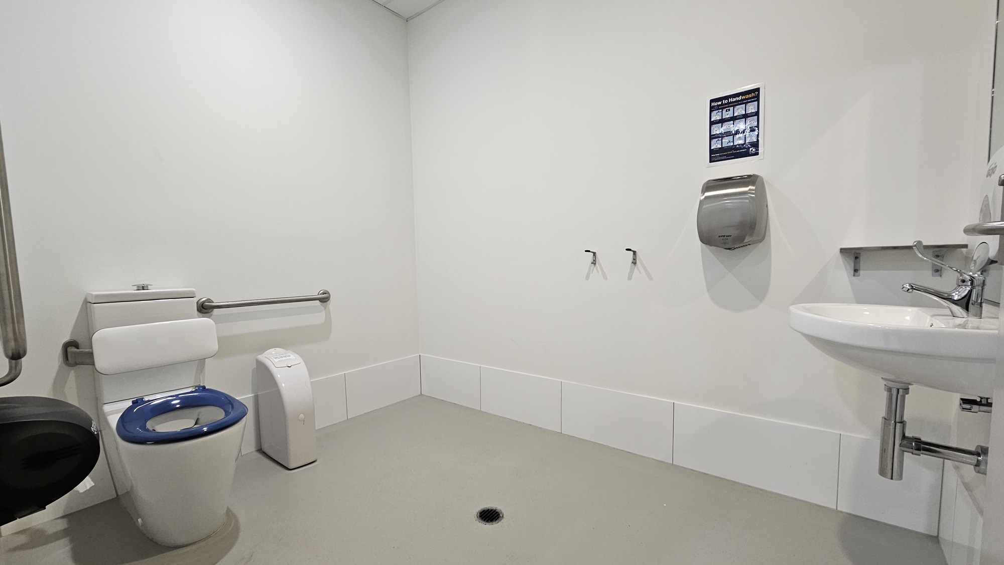 On the left, there is a toilet with a blue seat and a white cistern, accompanied by a grab rail on the wall. Next to the toilet is a white sanitary bin. The middle section of the wall is bare, with two hooks mounted on it. The right side features a wall-mounted stainless steel hand dryer and a white sink with a tap and soap dispenser. The floor is grey with a central drain, and the ceiling is white with recessed lighting. The background includes plain white walls.