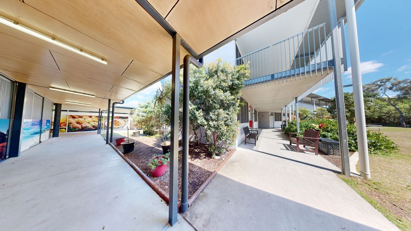 On the left, there's a two-storey building with a balcony containing several doors leading to rooms, alongside a wooden bench. The floor is grass, which is largely uniform and green. On the right, another section of the building is visible, with some greenery and trees nearby. There are picnic tables set on the grass, suggesting an outdoor area for gatherings. The ceiling is not visible, but the structure is modern with a flat roof. On the left, there's a two-storey building with a balcony containing several doors leading to rooms, alongside a wooden bench. The floor is grass, which is largely uniform and green. On the right, another section of the building is visible, with some greenery and trees nearby. There are picnic tables set on the grass, suggesting an outdoor area for gatherings. The ceiling is not visible, but the structure is modern with a flat roof.