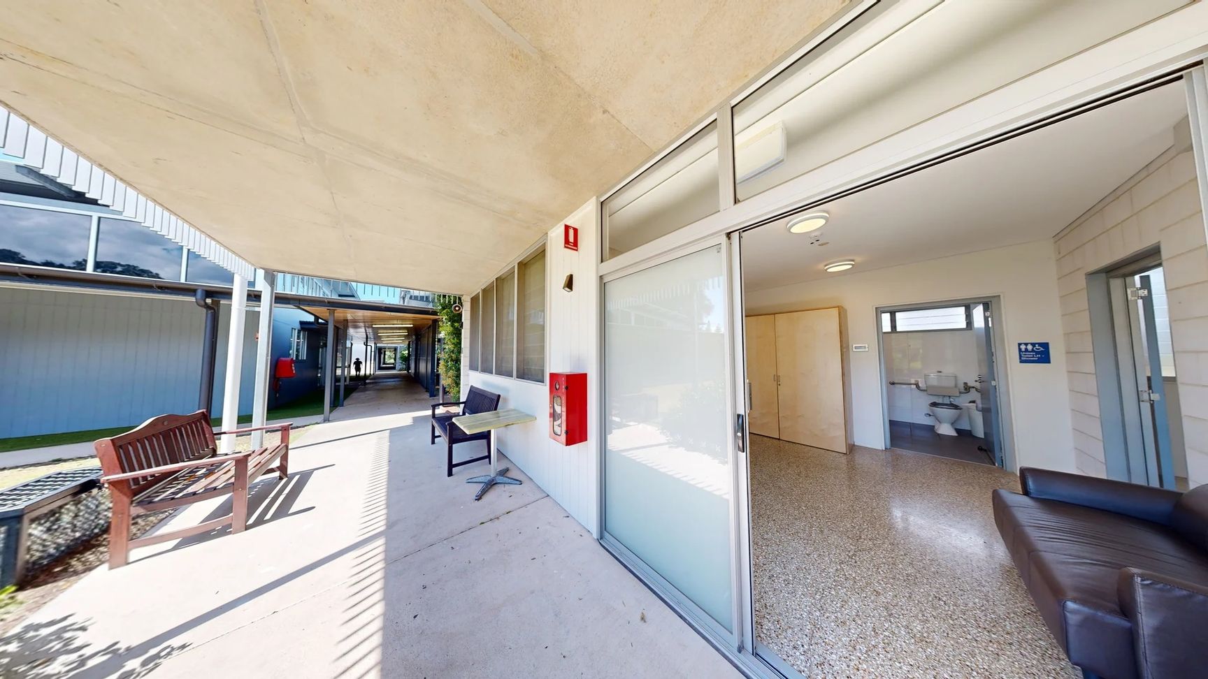 On the left, there is a covered walkway with a bench made of dark wood and a chair beside a small table. The floor is a smooth concrete surface, leading towards an open door revealing a room with light-coloured walls and a polished, light-coloured floor. To the right, there is a doorway leading into a bathroom space, showing a glimpse of fixtures. The ceiling is flat and has simple lighting fixtures arranged evenly. On the left, there is a covered walkway with a bench made of dark wood and a chair beside a small table. The floor is a smooth concrete surface, leading towards an open door revealing a room with light-coloured walls and a polished, light-coloured floor. To the right, there is a doorway leading into a bathroom space, showing a glimpse of fixtures. The ceiling is flat and has simple lighting fixtures arranged evenly.