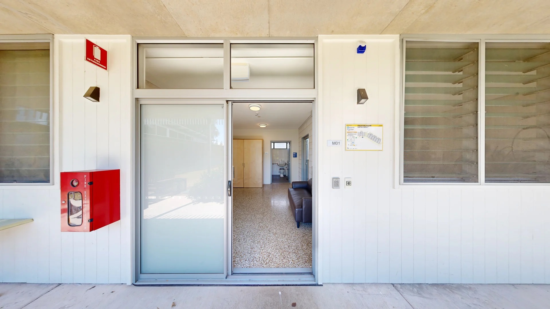 To the left, there is a bright red fire extinguisher box mounted on the wall, and beside it, a small, flat ledge. The entrance features a glass door leading into a well-lit interior with a light-coloured tiled floor. On the right, a series of large windows allow natural light inside, while a wall-mounted light fixture is positioned near the entrance. Inside, a sofa is visible in the foreground, facing an open doorway that leads to a bathroom in the background. To the left, there is a bright red fire extinguisher box mounted on the wall, and beside it, a small, flat ledge. The entrance features a glass door leading into a well-lit interior with a light-coloured tiled floor. On the right, a series of large windows allow natural light inside, while a wall-mounted light fixture is positioned near the entrance. Inside, a sofa is visible in the foreground, facing an open doorway that leads to a bathroom in the background.
