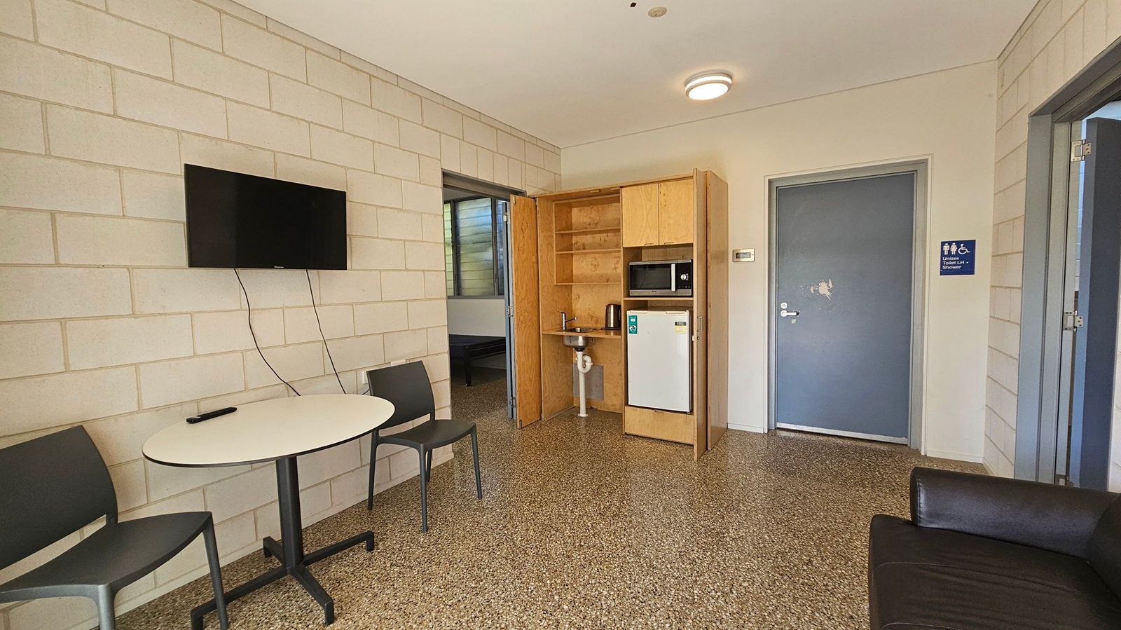 A room with light-coloured brick walls and a speckled floor. On the left, a wall-mounted TV is above a small round table with two chairs. In the centre, a wooden kitchenette features a sink, microwave, and mini-fridge. To the right, a blue door with a sign indicating a unisex accessible toilet. A black sofa is partially visible in the foreground. The ceiling is plain with a circular light fixture. A room with light-coloured brick walls and a speckled floor. On the left, a wall-mounted TV is above a small round table with two chairs. In the centre, a wooden kitchenette features a sink, microwave, and mini-fridge. To the right, a blue door with a sign indicating a unisex accessible toilet. A black sofa is partially visible in the foreground. The ceiling is plain with a circular light fixture.