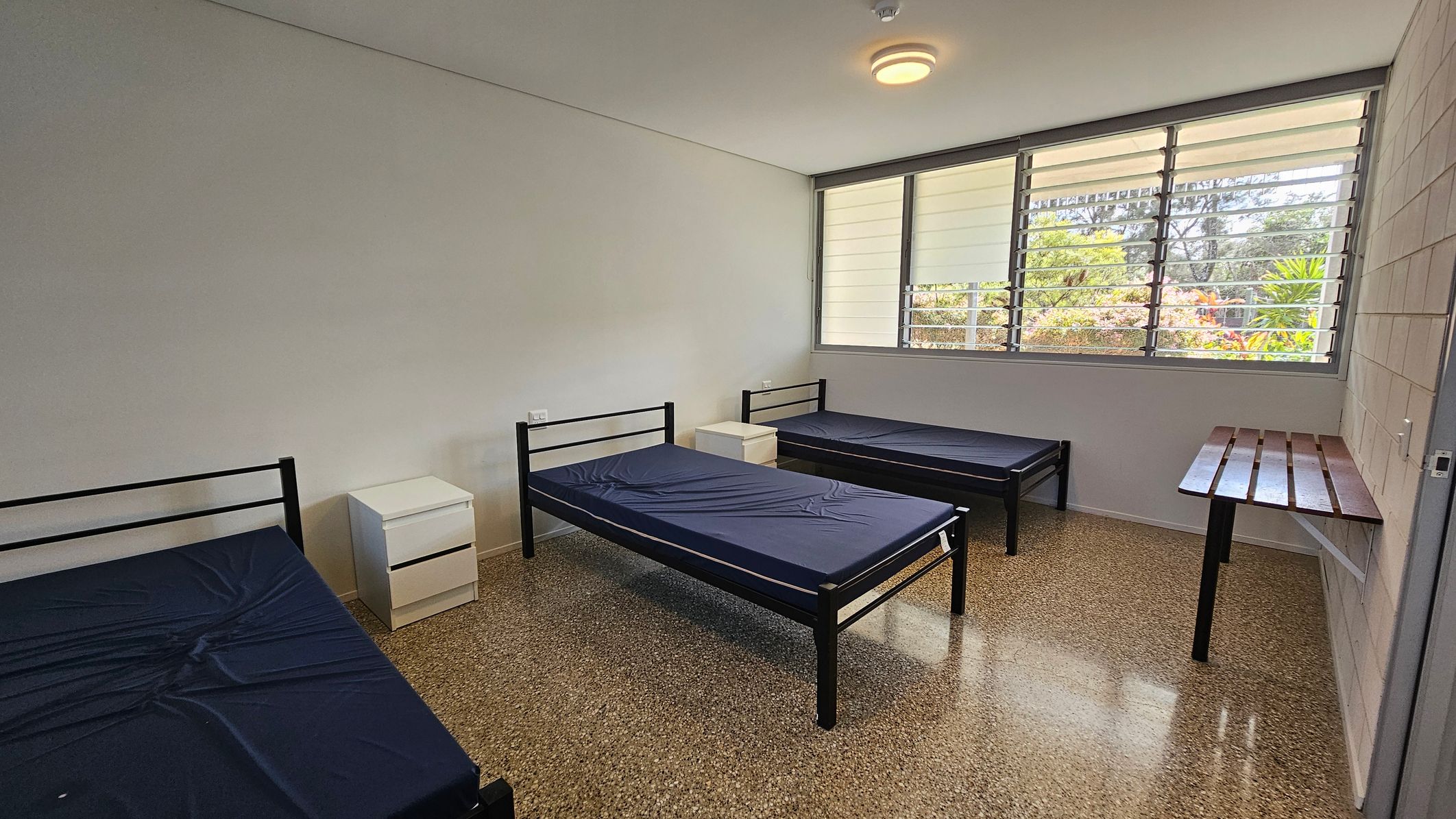 A room with three single metal-framed beds, each with a dark mattress. Two small white bedside tables are positioned between the beds. A large window with horizontal slats is on the right wall, allowing natural light to enter. Below the window is a simple wooden desk attached to the wall. The floor is speckled terrazzo, and the ceiling is plain with a central light fixture. A room with three single metal-framed beds, each with a dark mattress. Two small white bedside tables are positioned between the beds. A large window with horizontal slats is on the right wall, allowing natural light to enter. Below the window is a simple wooden desk attached to the wall. The floor is speckled terrazzo, and the ceiling is plain with a central light fixture.