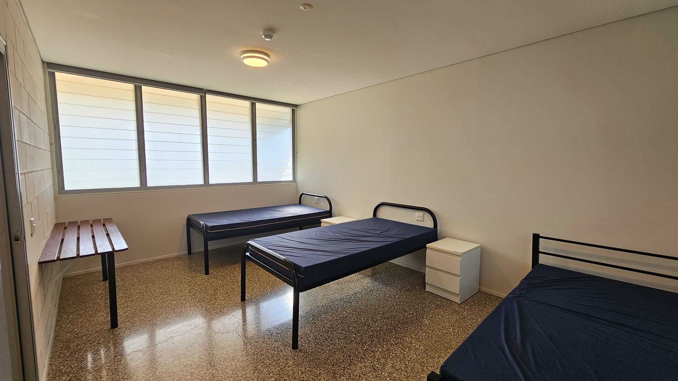 On the left, a wooden shelf rests against the wall, supported by two legs, providing space for personal items. There is a single bed with a dark blue mattress next to it. In the centre, another bed with a similar dark blue mattress occupies the middle of the room, framed by simple metal bed frames. On the right, a small white bedside table with drawers is positioned next to a third bed. The floor is a polished concrete with a smooth finish, and the ceiling is flat with a central light fixture. On the left, a wooden shelf rests against the wall, supported by two legs, providing space for personal items. There is a single bed with a dark blue mattress next to it. In the centre, another bed with a similar dark blue mattress occupies the middle of the room, framed by simple metal bed frames. On the right, a small white bedside table with drawers is positioned next to a third bed. The floor is a polished concrete with a smooth finish, and the ceiling is flat with a central light fixture.