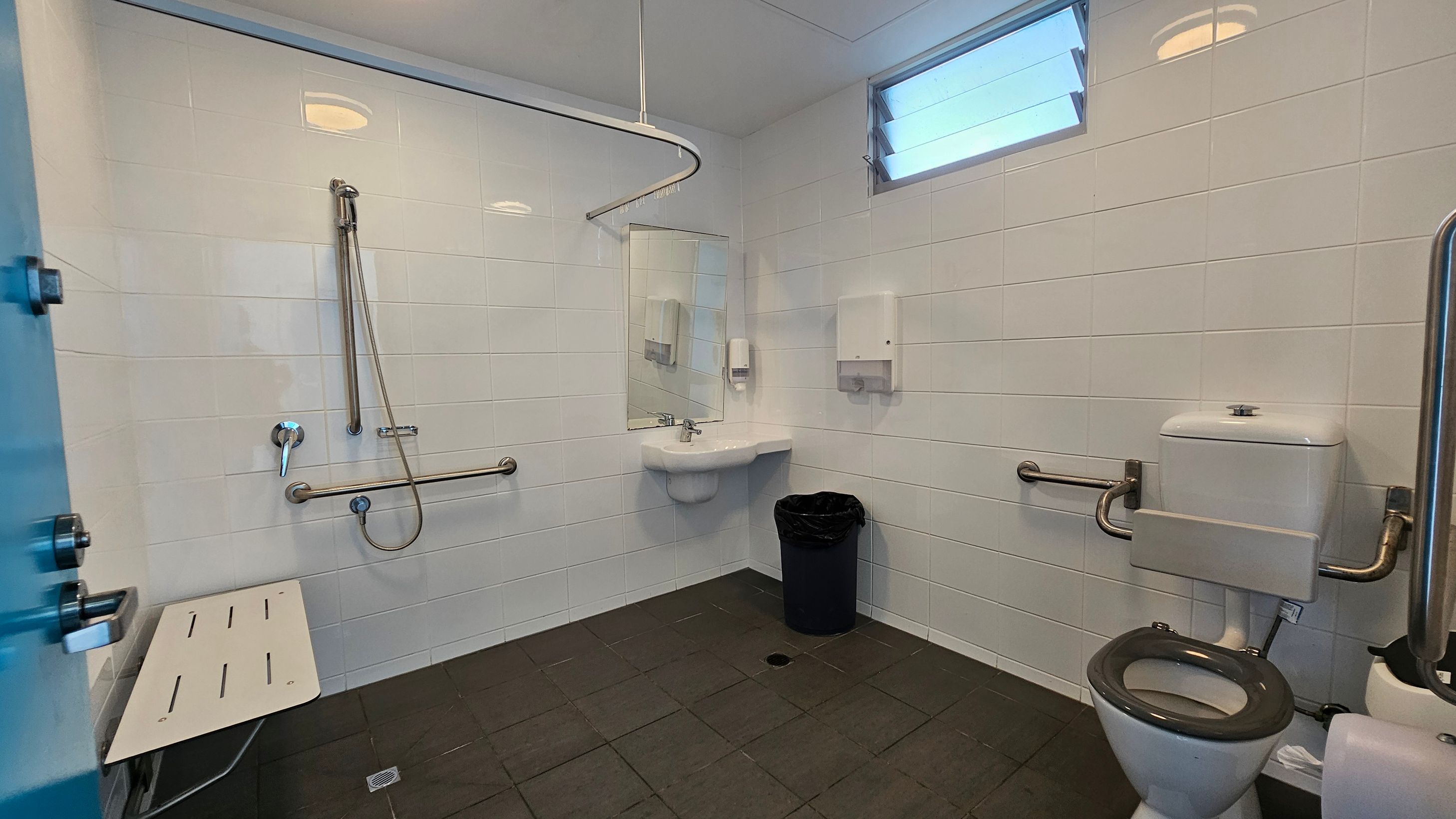 On the left, there is a blue door leading into the restroom, next to a vertical grab bar and a white folding shower seat mounted on the wall. To the right, a toilet is positioned against the wall, equipped with grab bars for assistance. In the centre, a small sink is mounted below a mirror, with a soap dispenser and paper towel holder nearby. The floor is covered in dark tiles, and the white walls have a glossy finish, reflecting light from overhead fixtures and a small window near the top. On the left, there is a blue door leading into the restroom, next to a vertical grab bar and a white folding shower seat mounted on the wall. To the right, a toilet is positioned against the wall, equipped with grab bars for assistance. In the centre, a small sink is mounted below a mirror, with a soap dispenser and paper towel holder nearby. The floor is covered in dark tiles, and the white walls have a glossy finish, reflecting light from overhead fixtures and a small window near the top.