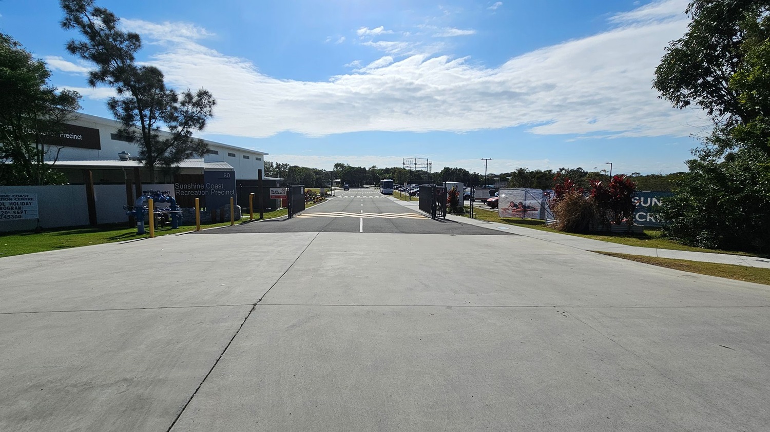 A smooth, concrete pathway stretches down the centre, flanked by greenery on the left and various structures on the right. To the left, there is a low building partially obscured by trees, with signage indicating the Sunshine Coast Recreation Precinct. On the right, a closed gate leads to a parking area, with cars visible in the distance. Beyond the pathway, the background features a few tall poles and additional vegetation, while the foreground showcases the well-maintained concrete surface A smooth, concrete pathway stretches down the centre, flanked by greenery on the left and various structures on the right. To the left, there is a low building partially obscured by trees, with signage indicating the Sunshine Coast Recreation Precinct. On the right, a closed gate leads to a parking area, with cars visible in the distance. Beyond the pathway, the background features a few tall poles and additional vegetation, while the foreground showcases the well-maintained concrete surface