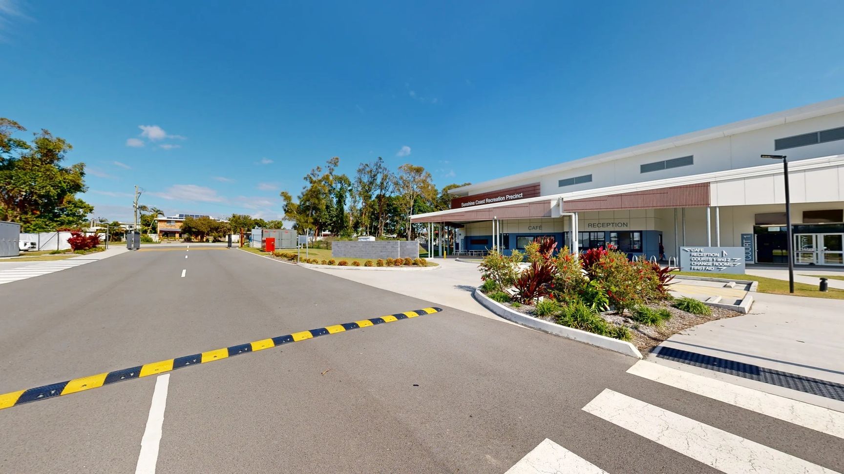 A wide grey asphalt road extends towards a building in the centre. On the left, a landscaped area features various plants and a path leading to an entrance. A red postal box stands prominently. To the right, the modern structure has large glass windows, with visible signage for a café and reception. Brightly coloured foliage outlines the entrance, and a smooth pedestrian crossing indicates safe passage. A wide grey asphalt road extends towards a building in the centre. On the left, a landscaped area features various plants and a path leading to an entrance. A red postal box stands prominently. To the right, the modern structure has large glass windows, with visible signage for a café and reception. Brightly coloured foliage outlines the entrance, and a smooth pedestrian crossing indicates safe passage.