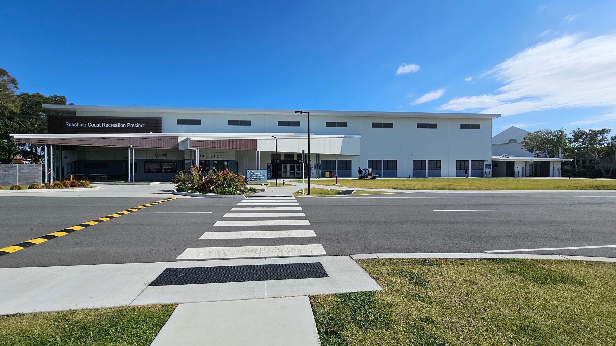 A wide, smooth grey road leads directly to a large white building in the centre, labelled "Sunshine Coast Recreation Precinct." On the left, there is a covered area with outdoor seating and vibrant landscaping. To the right, additional sections of the building include a reception area and an entrance with automatic doors. The foreground features a wide, marked pedestrian crossing with textured pavers, while the background includes patches of grass and trees bordering the area. A wide, smooth grey road leads directly to a large white building in the centre, labelled "Sunshine Coast Recreation Precinct." On the left, there is a covered area with outdoor seating and vibrant landscaping. To the right, additional sections of the building include a reception area and an entrance with automatic doors. The foreground features a wide, marked pedestrian crossing with textured pavers, while the background includes patches of grass and trees bordering the area.