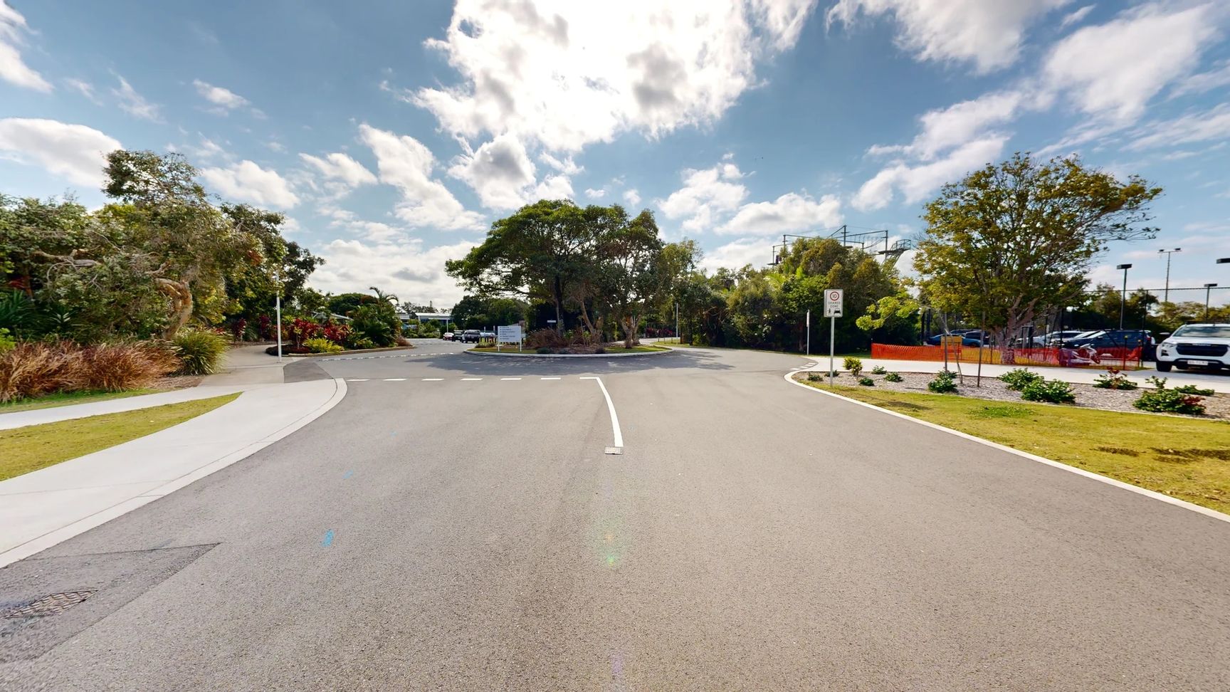 A smooth road curves gently ahead, leading into a roundabout. On the left, a well-kept landscape features various shrubs and small trees, with a paved walkway alongside. To the right, there is a parking area, partially obscured by greenery, and several vehicles are parked in view. In the foreground, the clean road surface is clearly marked with a white line indicating the curve. The background includes more dense foliage, with a large tree providing shade near the centre of the roundabout. A smooth road curves gently ahead, leading into a roundabout. On the left, a well-kept landscape features various shrubs and small trees, with a paved walkway alongside. To the right, there is a parking area, partially obscured by greenery, and several vehicles are parked in view. In the foreground, the clean road surface is clearly marked with a white line indicating the curve. The background includes more dense foliage, with a large tree providing shade near the centre of the roundabout.