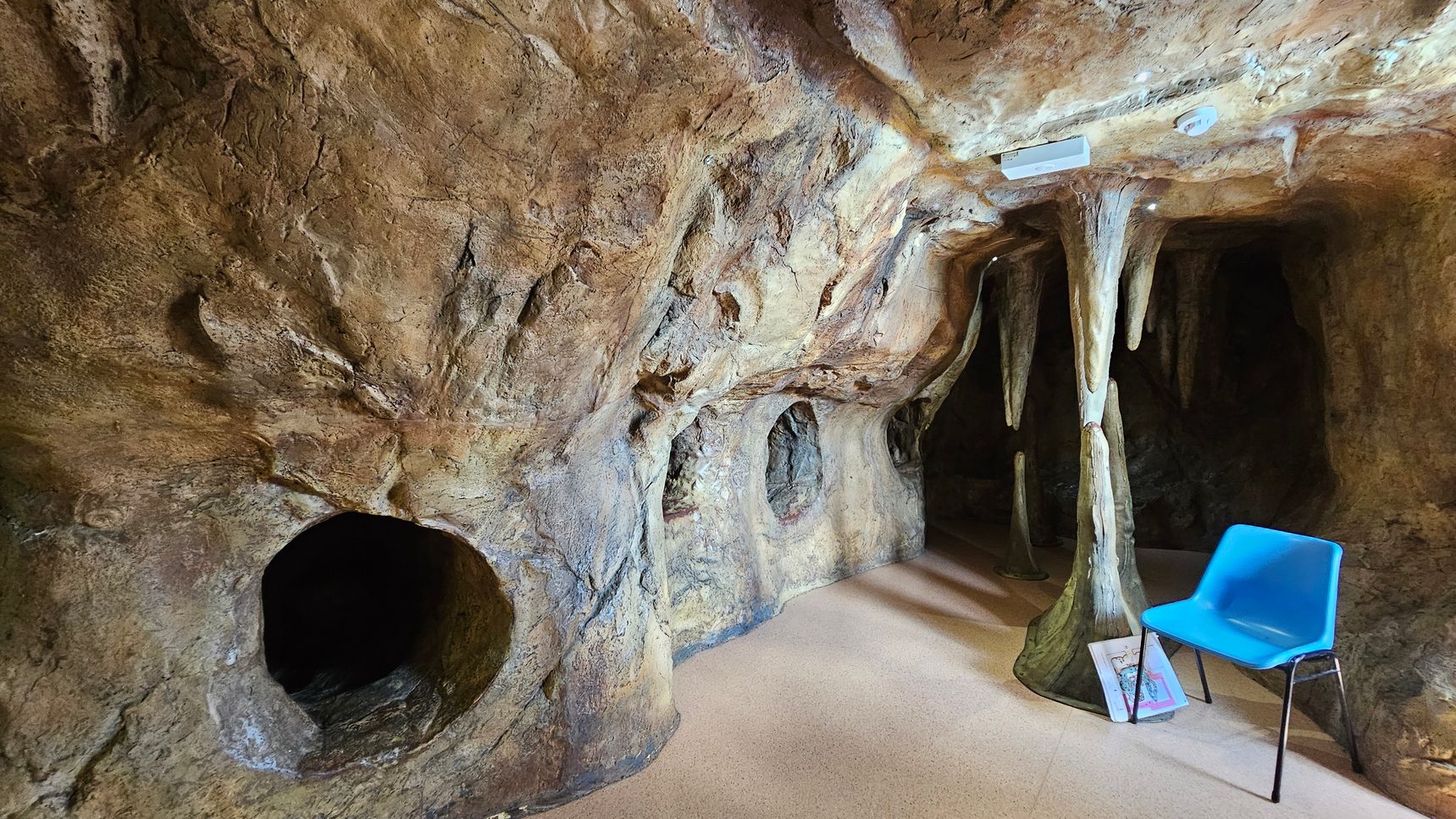 A cave-like room with textured, rocky walls and ceiling. On the right, a blue plastic chair sits on a beige floor, next to a stack of books. Stalactite and stalagmite formations are present, with one column reaching from floor to ceiling. The left side features circular openings in the rock wall, leading to dark recesses. The lighting is dim, enhancing the cave atmosphere. A cave-like room with textured, rocky walls and ceiling. On the right, a blue plastic chair sits on a beige floor, next to a stack of books. Stalactite and stalagmite formations are present, with one column reaching from floor to ceiling. The left side features circular openings in the rock wall, leading to dark recesses. The lighting is dim, enhancing the cave atmosphere.