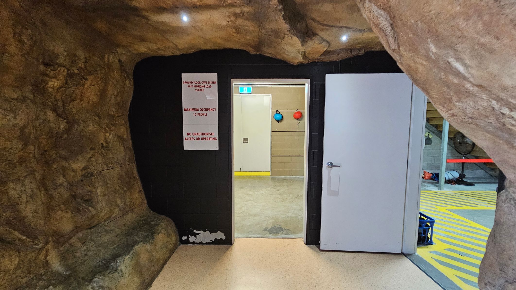 A cave-like interior with rough, rocky walls and ceiling. In the centre, a white door is open, leading to a room with a concrete floor and beige walls. Two coloured buoys hang on the wall inside. A sign on the left of the door reads "Ground Floor Cave System Safe Working Load 1500kg, Maximum Occupancy 15 People, No Unauthorised Access or Operating." The floor is light-coloured, and the ceiling has small recessed lights. A cave-like interior with rough, rocky walls and ceiling. In the centre, a white door is open, leading to a room with a concrete floor and beige walls. Two coloured buoys hang on the wall inside. A sign on the left of the door reads "Ground Floor Cave System Safe Working Load 1500kg, Maximum Occupancy 15 People, No Unauthorised Access or Operating." The floor is light-coloured, and the ceiling has small recessed lights.