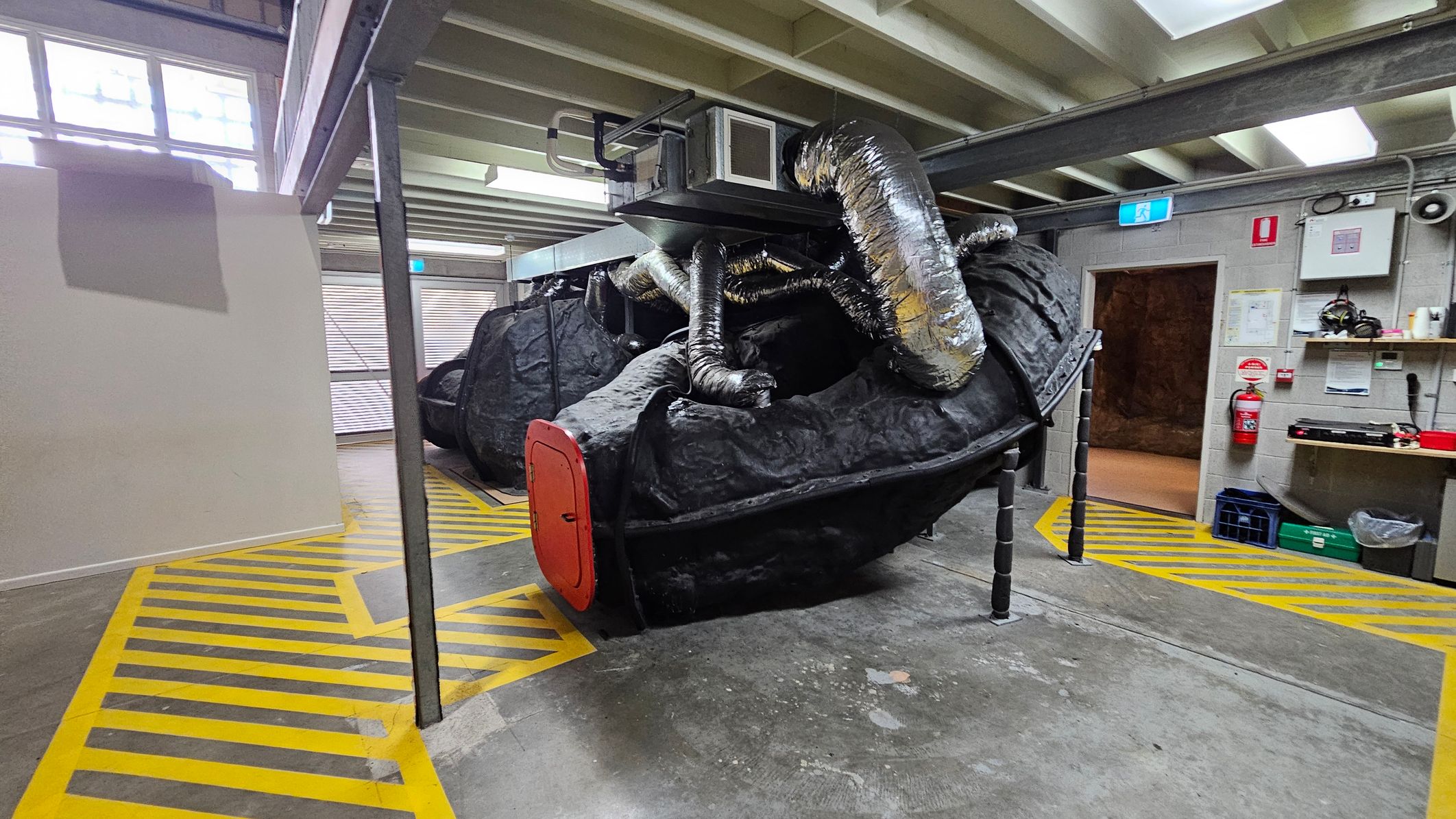 A large, black, rock-like structure with a red door is in the centre, supported by metal legs. Silver ducting tubes are attached to the top, connected to a ventilation unit. The floor has yellow and black striped markings. On the right, there's a wall with a fire extinguisher, safety signs, and shelves holding various items. The ceiling is made of exposed beams with fluorescent lights. Windows on the left let in natural light. A large, black, rock-like structure with a red door is in the centre, supported by metal legs. Silver ducting tubes are attached to the top, connected to a ventilation unit. The floor has yellow and black striped markings. On the right, there's a wall with a fire extinguisher, safety signs, and shelves holding various items. The ceiling is made of exposed beams with fluorescent lights. Windows on the left let in natural light.