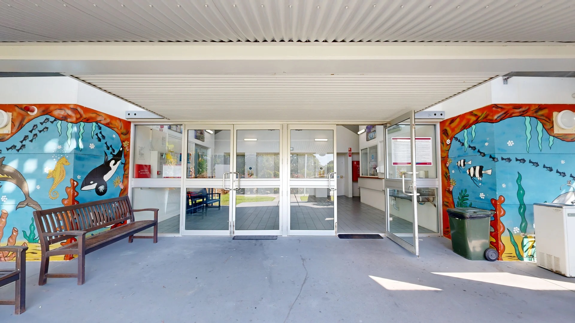 On the left, a sign provides information about the facility, mounted on a white pole, with a colourful mural of sea life visible beside it. To the right, a large rock with a sign labelled “Caves Rock Face” sits near vibrant tropical plants. The pathway is made of smooth grey concrete, leading towards double glass doors. The ceiling above is gently sloped, with a large overhang sheltering the entrance. The surrounding area features well-maintained grass and various tropical foliage. On the left, a sign provides information about the facility, mounted on a white pole, with a colourful mural of sea life visible beside it. To the right, a large rock with a sign labelled “Caves Rock Face” sits near vibrant tropical plants. The pathway is made of smooth grey concrete, leading towards double glass doors. The ceiling above is gently sloped, with a large overhang sheltering the entrance. The surrounding area features well-maintained grass and various tropical foliage.