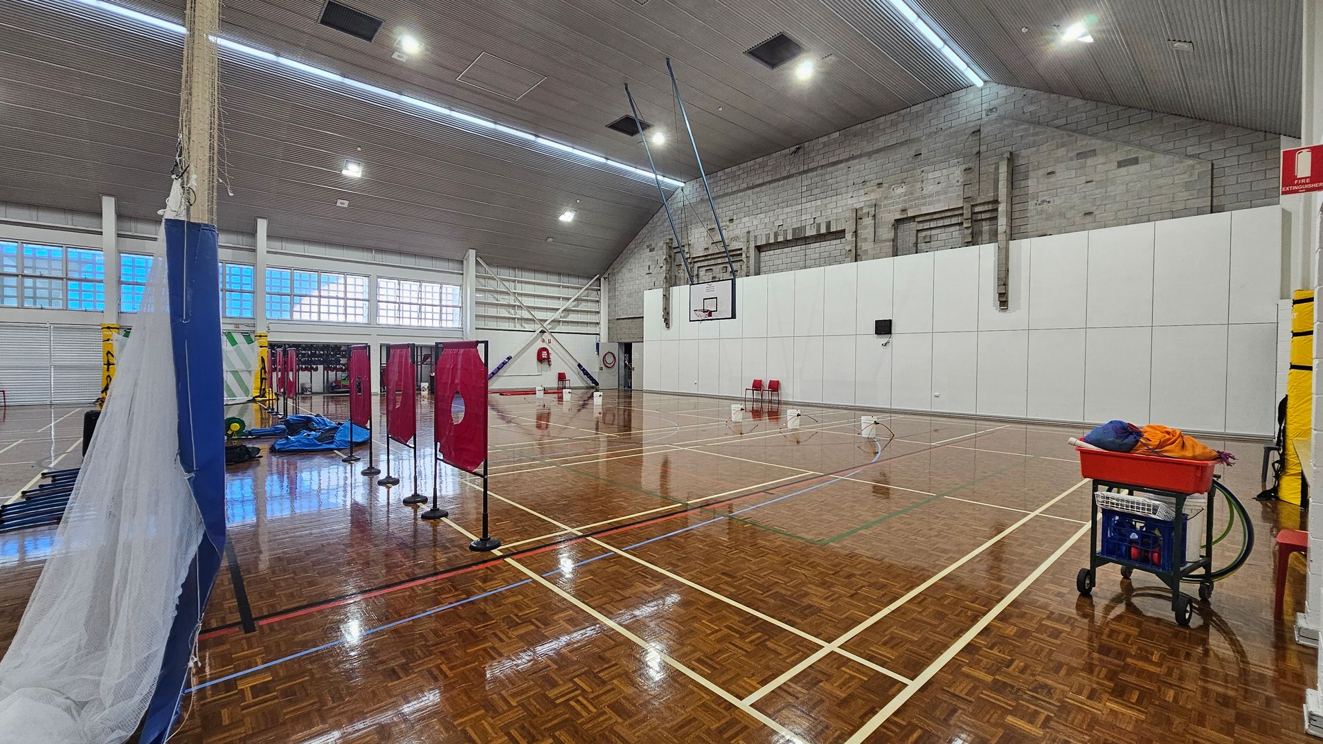 On the left, a tall blue structure is partially covered by a net, with various sports equipment scattered nearby, including a blue tarp. Red targets on stands are lined up in the centre. To the right, a white wall features a basketball hoop, with two red chairs placed beneath it. The floor is polished wood with visible lines indicating court boundaries. The ceiling is high and features a textured design, with lights evenly distributed across its surface. On the left, a tall blue structure is partially covered by a net, with various sports equipment scattered nearby, including a blue tarp. Red targets on stands are lined up in the centre. To the right, a white wall features a basketball hoop, with two red chairs placed beneath it. The floor is polished wood with visible lines indicating court boundaries. The ceiling is high and features a textured design, with lights evenly distributed across its surface.