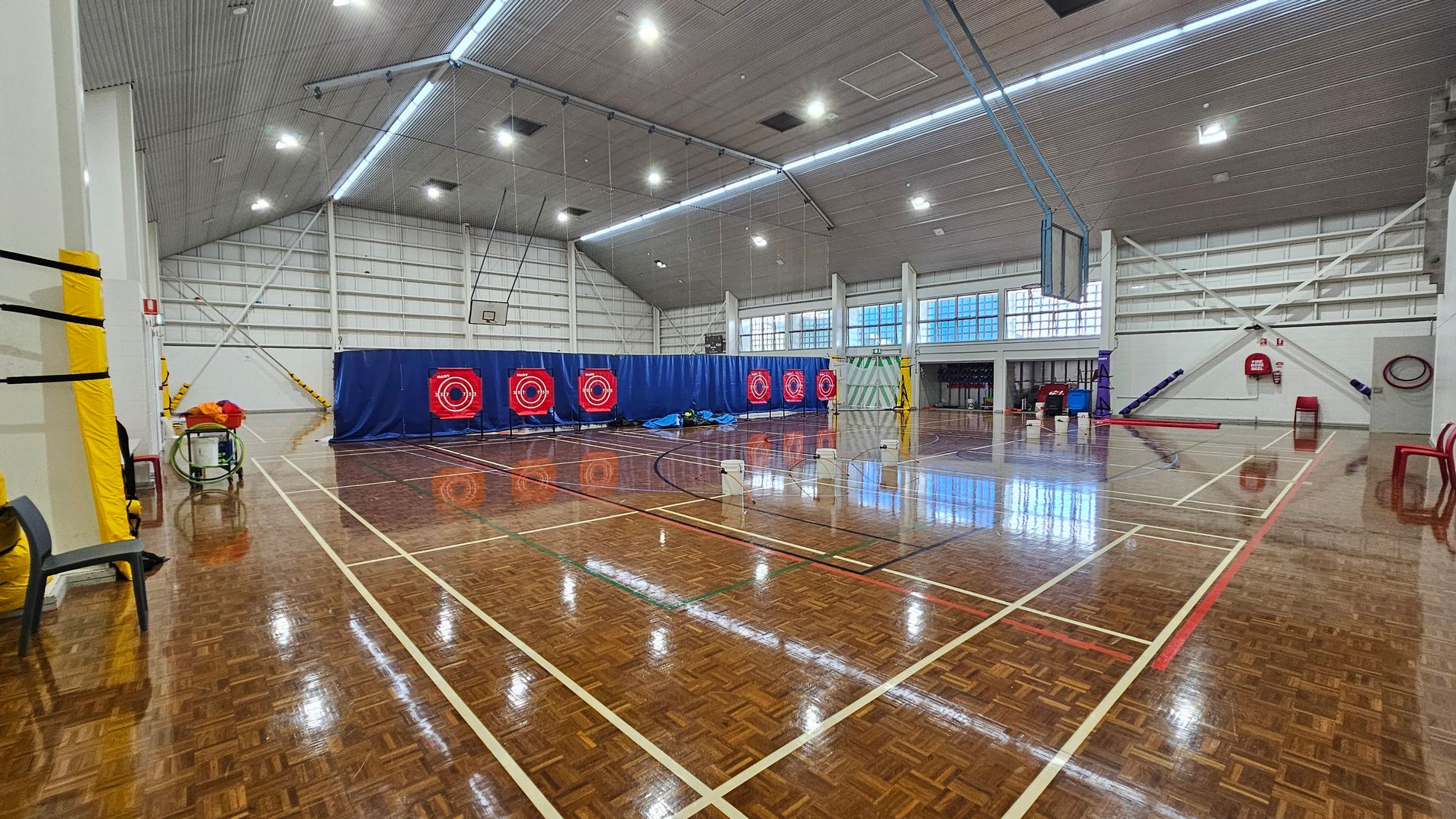 On the left, a tall wall has basketball hoops attached, with a red padded area below. Nearby are various colourful mats stacked against the wall, providing cushioning for activities. To the right, a scoreboard is mounted, along with large windows that allow natural light into the space. The polished wooden floor features court markings in white and green, extending across the area. The ceiling is sloped with bright lighting evenly distributed to illuminate the gymnasium effectively. On the left, a tall wall has basketball hoops attached, with a red padded area below. Nearby are various colourful mats stacked against the wall, providing cushioning for activities. To the right, a scoreboard is mounted, along with large windows that allow natural light into the space. The polished wooden floor features court markings in white and green, extending across the area. The ceiling is sloped with bright lighting evenly distributed to illuminate the gymnasium effectively.