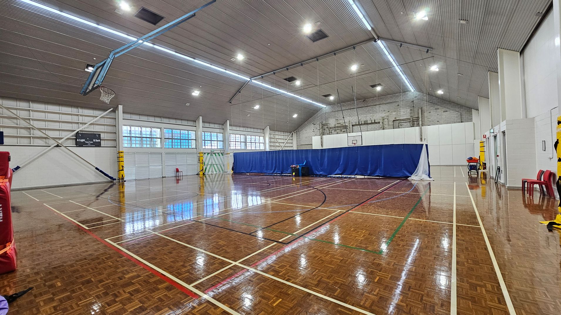 On the left, a red padded wall is partially visible, with a basketball hoop fixed above it, and large windows allowing light into the space. To the right, a blue curtain separates part of the area, with a couple of red chairs positioned nearby. The glossy wooden floor features white and coloured lines marking different court boundaries. The ceiling is high with a ribbed texture, fitted with bright overhead lights that provide ample illumination throughout the gymnasium. On the left, a red padded wall is partially visible, with a basketball hoop fixed above it, and large windows allowing light into the space. To the right, a blue curtain separates part of the area, with a couple of red chairs positioned nearby. The glossy wooden floor features white and coloured lines marking different court boundaries. The ceiling is high with a ribbed texture, fitted with bright overhead lights that provide ample illumination throughout the gymnasium.