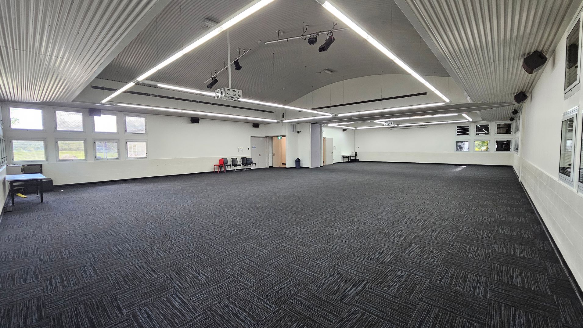 On the left, a small table with a blue cloth is set against the wall, accompanied by a few grey chairs. To the right, a doorway leads to another room, and there are additional chairs positioned nearby. The floor is carpeted in a dark pattern, and the spacious area is largely empty. The ceiling is curved with a corrugated design, featuring bright LED lights running across it, providing even illumination throughout the room. On the left, a small table with a blue cloth is set against the wall, accompanied by a few grey chairs. To the right, a doorway leads to another room, and there are additional chairs positioned nearby. The floor is carpeted in a dark pattern, and the spacious area is largely empty. The ceiling is curved with a corrugated design, featuring bright LED lights running across it, providing even illumination throughout the room.