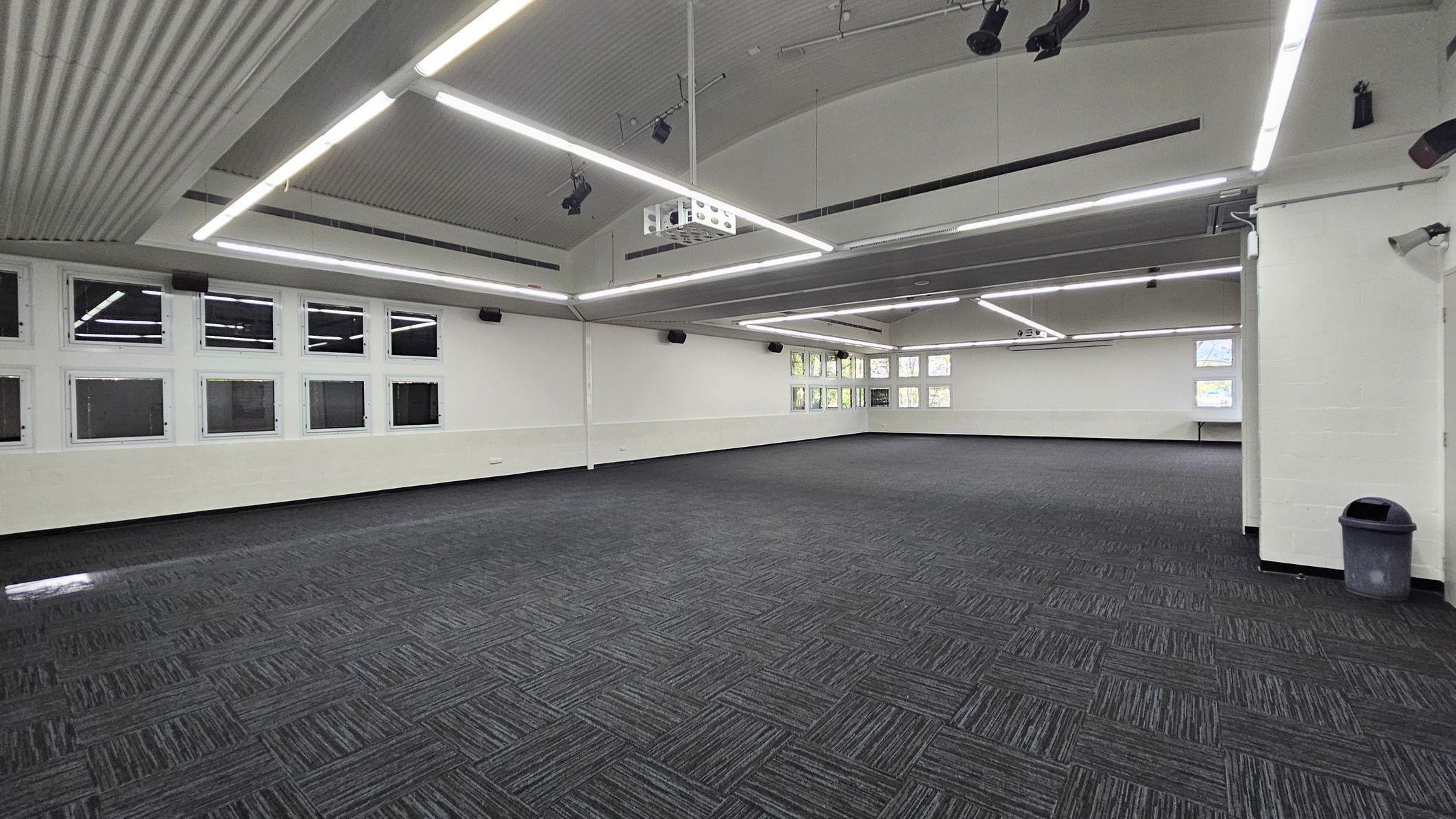 On the left, a row of small windows is situated just above a low white wall, allowing light to enter the room. To the right, a grey rubbish bin is positioned next to a small corner area with a low countertop that is unobtrusive. The floor is covered in dark, textured carpeting laid in a patterned design. The ceiling arches gently and features several bright, linear lights along its length, illuminating the spacious interior. On the left, a row of small windows is situated just above a low white wall, allowing light to enter the room. To the right, a grey rubbish bin is positioned next to a small corner area with a low countertop that is unobtrusive. The floor is covered in dark, textured carpeting laid in a patterned design. The ceiling arches gently and features several bright, linear lights along its length, illuminating the spacious interior.