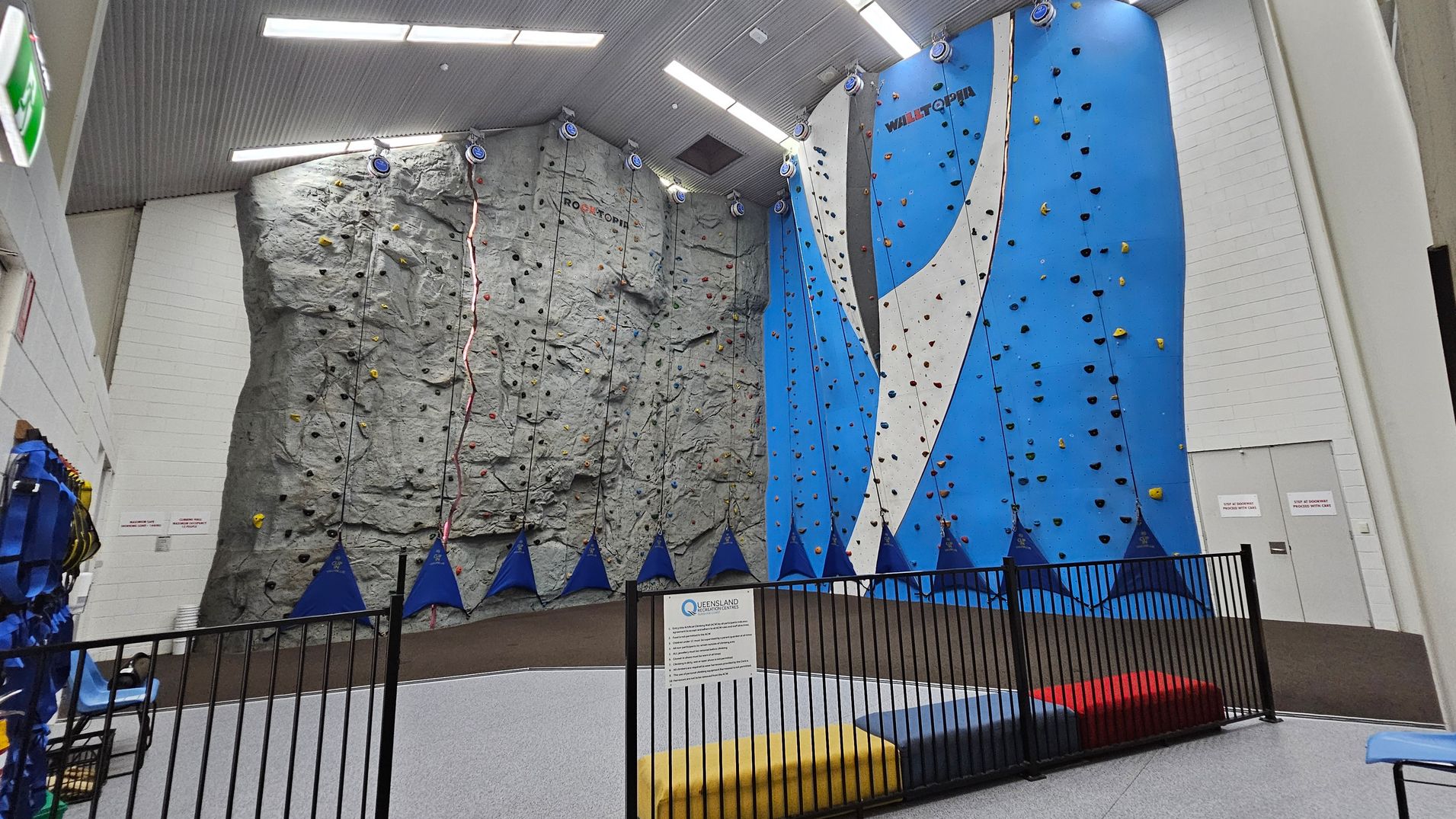 Double doors are open, revealing a soft, grey floor and a low wall with a safety railing on the left. A vibrant red mat is positioned in the centre, with blue climbing shapes in the background, hinting at climbing activities. To the right, there's a wall decorated with the words "The Rock Face" in bold, modern typography, accompanied by a geometric blue design. The ceiling is plain and light-coloured, providing a subtle contrast to the walls. Double doors are open, revealing a soft, grey floor and a low wall with a safety railing on the left. A vibrant red mat is positioned in the centre, with blue climbing shapes in the background, hinting at climbing activities. To the right, there's a wall decorated with the words "The Rock Face" in bold, modern typography, accompanied by a geometric blue design. The ceiling is plain and light-coloured, providing a subtle contrast to the walls.