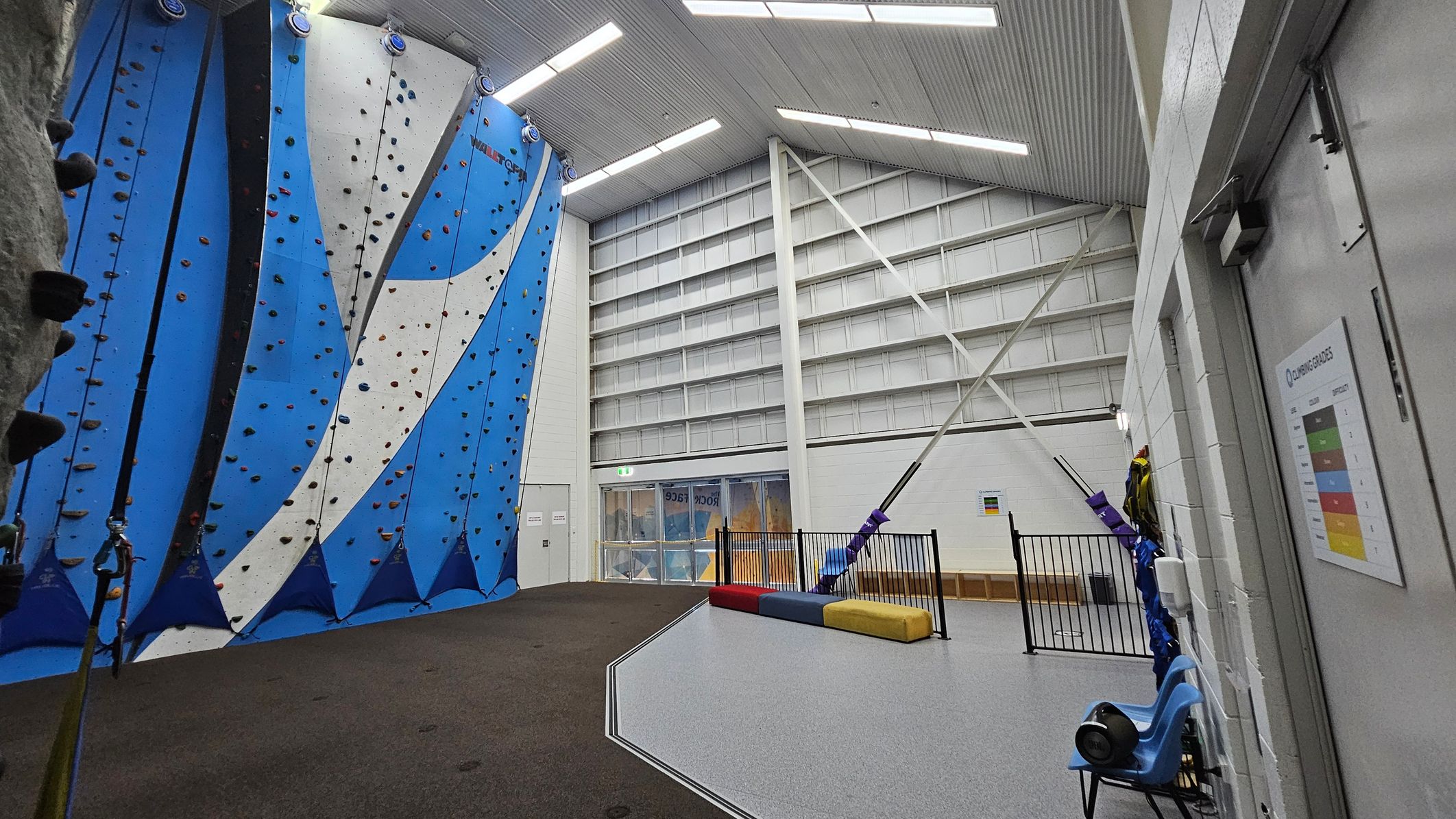 Indoor climbing gym with blue and white climbing walls on the left, featuring various holds. The floor is a mix of dark and light grey surfaces. On the right, a white wall with a climbing grades chart and a blue chair. In the background, a large windowed exit door and a fenced-off area with colourful padded blocks. The ceiling is high with exposed beams and fluorescent lights. Indoor climbing gym with blue and white climbing walls on the left, featuring various holds. The floor is a mix of dark and light grey surfaces. On the right, a white wall with a climbing grades chart and a blue chair. In the background, a large windowed exit door and a fenced-off area with colourful padded blocks. The ceiling is high with exposed beams and fluorescent lights.