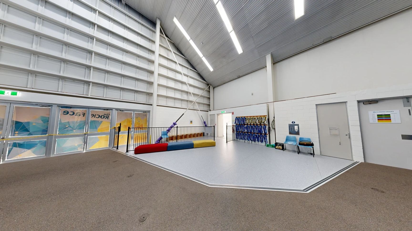 On the left, large double doors with patterned glass reveal a vibrant, geometric design behind them. A railing surrounds a colourful mat in red, yellow, and blue, positioned in the centre of the space. To the right, a wall displays several rows of blue harnesses, and two light blue chairs are placed nearby. The floor has a mix of light grey and soft brown textures. The ceiling is high with a series of linear lights running centrally, and is lined with corrugated metal. On the left, large double doors with patterned glass reveal a vibrant, geometric design behind them. A railing surrounds a colourful mat in red, yellow, and blue, positioned in the centre of the space. To the right, a wall displays several rows of blue harnesses, and two light blue chairs are placed nearby. The floor has a mix of light grey and soft brown textures. The ceiling is high with a series of linear lights running centrally, and is lined with corrugated metal.