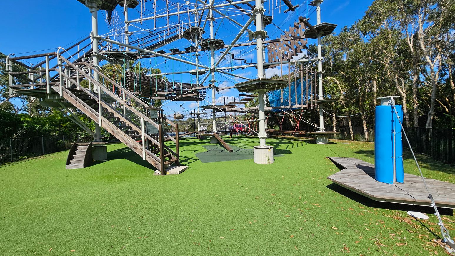 On the left, there are metal bleachers with a white canopy above, supported by metal poles. In the centre, a large outdoor climbing structure with ropes and platforms is visible, labelled "Sunshine Coast." The ground is paved with concrete, leading to a grassy area. On the right, there are various green and red-leaved plants and trees, providing a natural backdrop. On the left, there are metal bleachers with a white canopy above, supported by metal poles. In the centre, a large outdoor climbing structure with ropes and platforms is visible, labelled "Sunshine Coast." The ground is paved with concrete, leading to a grassy area. On the right, there are various green and red-leaved plants and trees, providing a natural backdrop.