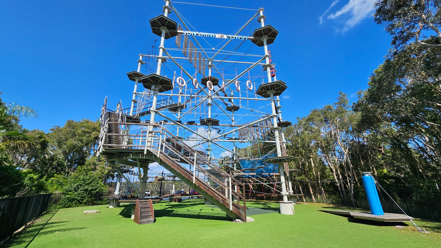 A large outdoor climbing structure with multiple levels and platforms, featuring ropes and obstacles. The structure is supported by metal poles and has a staircase on the left side. The ground is covered with artificial green grass. Surrounding the structure are tall trees and a clear blue sky. A blue cylindrical object is on the right side near the base. A large outdoor climbing structure with multiple levels and platforms, featuring ropes and obstacles. The structure is supported by metal poles and has a staircase on the left side. The ground is covered with artificial green grass. Surrounding the structure are tall trees and a clear blue sky. A blue cylindrical object is on the right side near the base.