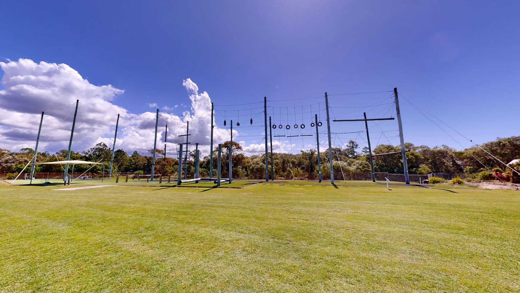 To the left, there is a large, green, rectangular area of grass, alongside a low structure that appears to be a support for a zip line or swinging apparatus. To the right, several vertical poles are arranged, some with horizontal bars and others adorned with tyres, indicating a challenge course. In the background, lush trees provide coverage, while the foreground is a well-kept expanse of grass. No notable details of the floor or ceiling are visible in this setting. To the left, there is a large, green, rectangular area of grass, alongside a low structure that appears to be a support for a zip line or swinging apparatus. To the right, several vertical poles are arranged, some with horizontal bars and others adorned with tyres, indicating a challenge course. In the background, lush trees provide coverage, while the foreground is a well-kept expanse of grass. No notable details of the floor or ceiling are visible in this setting.