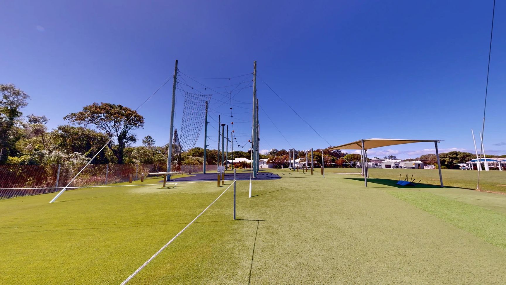 On the left, a series of tall poles are set up, connected by wires and nets, forming an obstacle course. A dark rubber flooring area is visible beneath them. On the right, there is a shaded structure with a sloping roof, supported by poles, and a blue mat or equipment situated beneath it. The ground is covered in even green artificial grass, extending across the scene. In the background, trees and low buildings are present, providing a natural setting. On the left, a series of tall poles are set up, connected by wires and nets, forming an obstacle course. A dark rubber flooring area is visible beneath them. On the right, there is a shaded structure with a sloping roof, supported by poles, and a blue mat or equipment situated beneath it. The ground is covered in even green artificial grass, extending across the scene. In the background, trees and low buildings are present, providing a natural setting.