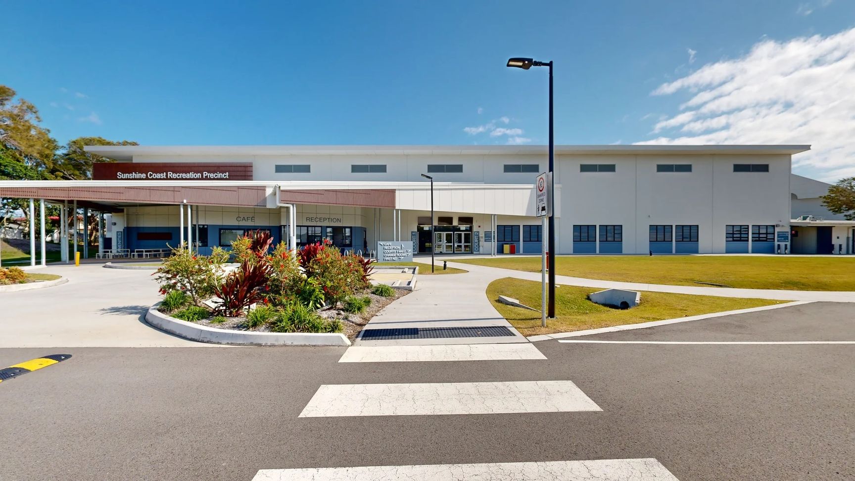 A wide, grey asphalt pathway leads directly to a large building marked "Sunshine Coast Recreation Precinct" at the centre. On the left, outdoor seating is positioned under a roofed area, alongside landscaped plant beds with colourful foliage. To the right, large glass doors lead to the reception and café areas, with signage visible above. The well-maintained concrete floor of the pathway blends into the surrounding grass, which is evenly cut. A wide, grey asphalt pathway leads directly to a large building marked "Sunshine Coast Recreation Precinct" at the centre. On the left, outdoor seating is positioned under a roofed area, alongside landscaped plant beds with colourful foliage. To the right, large glass doors lead to the reception and café areas, with signage visible above. The well-maintained concrete floor of the pathway blends into the surrounding grass, which is evenly cut.