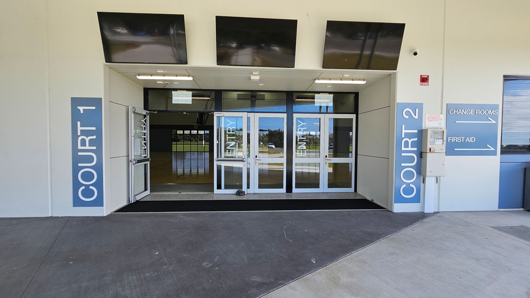 At the entrance, two sets of glass doors lead into a spacious interior. On the left, a large panel displays "COURT 1," while on the right, another panel indicates "COURT 2," alongside directional signs for "CHANGE ROOMS" and "FIRST AID." Above the doors, there are three dark ceiling panels, and fluorescent lights illuminate the entrance area. The ground is a smooth concrete surface with a slight slope towards the entrance, providing easy access. At the entrance, two sets of glass doors lead into a spacious interior. On the left, a large panel displays "COURT 1," while on the right, another panel indicates "COURT 2," alongside directional signs for "CHANGE ROOMS" and "FIRST AID." Above the doors, there are three dark ceiling panels, and fluorescent lights illuminate the entrance area. The ground is a smooth concrete surface with a slight slope towards the entrance, providing easy access.