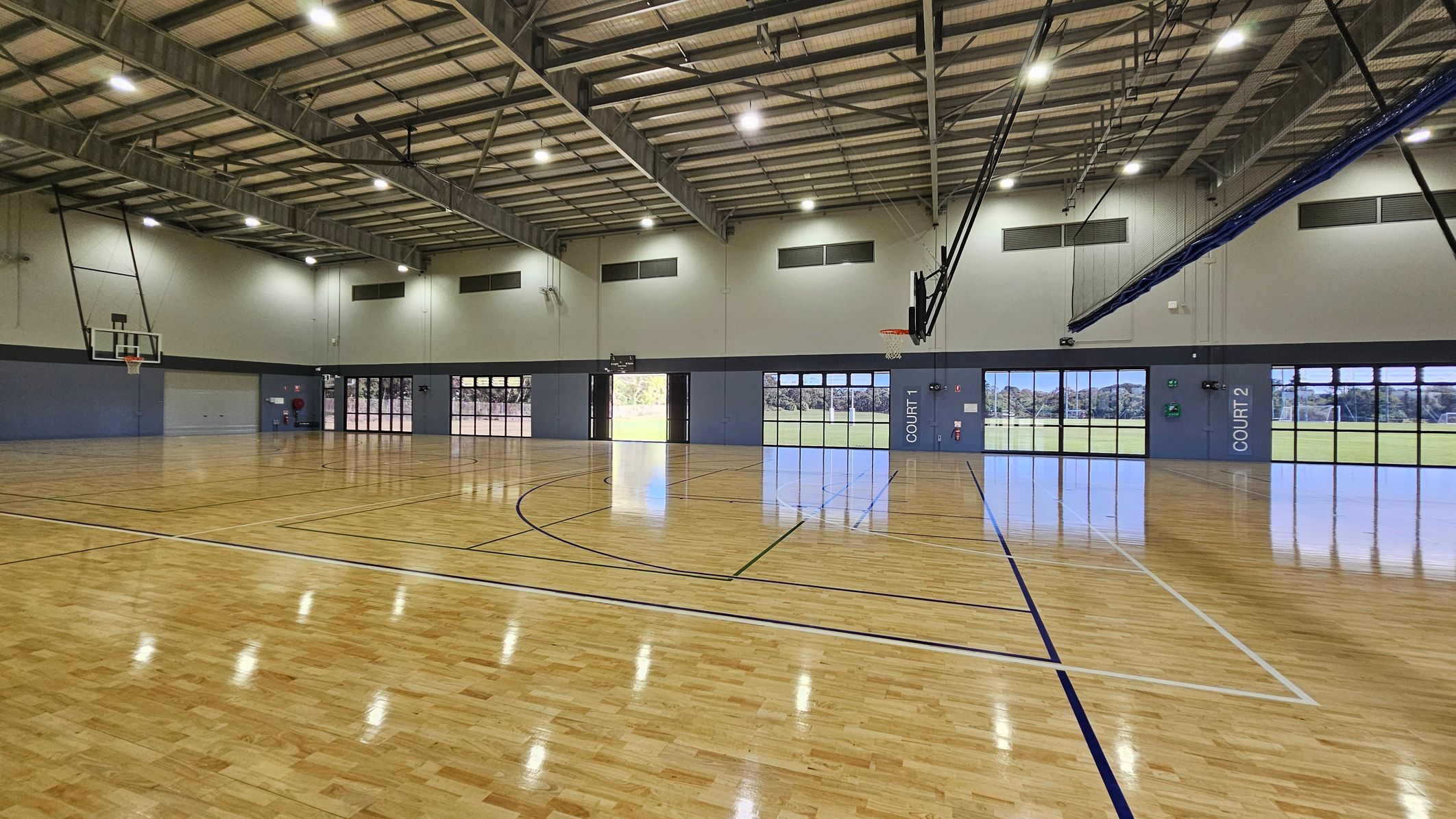 In a large indoor sports hall, polished wooden floors stretch across the area, reflecting the light from overhead fluorescent fixtures. On the left side, a basketball hoop hangs above a blue wall with an entrance door nearby. To the right, two large glass doors marked "COURT 2" reveal an outdoor field beyond. Each side features large windows allowing natural light into the space. The ceiling is high and lined with metal beams, creating an open atmosphere. In a large indoor sports hall, polished wooden floors stretch across the area, reflecting the light from overhead fluorescent fixtures. On the left side, a basketball hoop hangs above a blue wall with an entrance door nearby. To the right, two large glass doors marked "COURT 2" reveal an outdoor field beyond. Each side features large windows allowing natural light into the space. The ceiling is high and lined with metal beams, creating an open atmosphere.