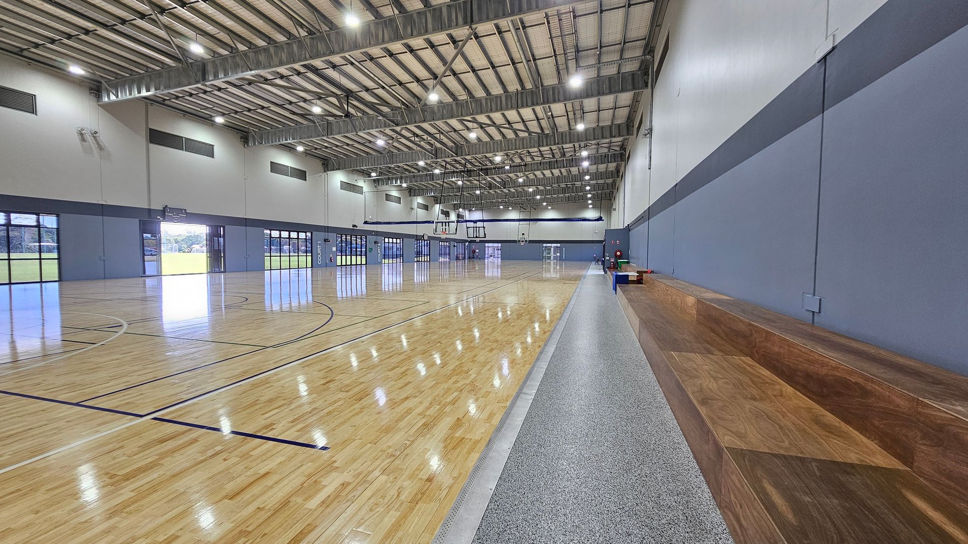 The sports hall features polished wooden flooring that reflects overhead lights, with court markings visible across the surface. On the left, a smooth concrete area runs parallel to the court, leading to a wooden bench that provides seating. To the right, a blue wall has a door and safety equipment visible, including a fire extinguisher. The ceiling is high, constructed with metal beams and lined with bright fluorescent lights that illuminate the space evenly. The sports hall features polished wooden flooring that reflects overhead lights, with court markings visible across the surface. On the left, a smooth concrete area runs parallel to the court, leading to a wooden bench that provides seating. To the right, a blue wall has a door and safety equipment visible, including a fire extinguisher. The ceiling is high, constructed with metal beams and lined with bright fluorescent lights that illuminate the space evenly.