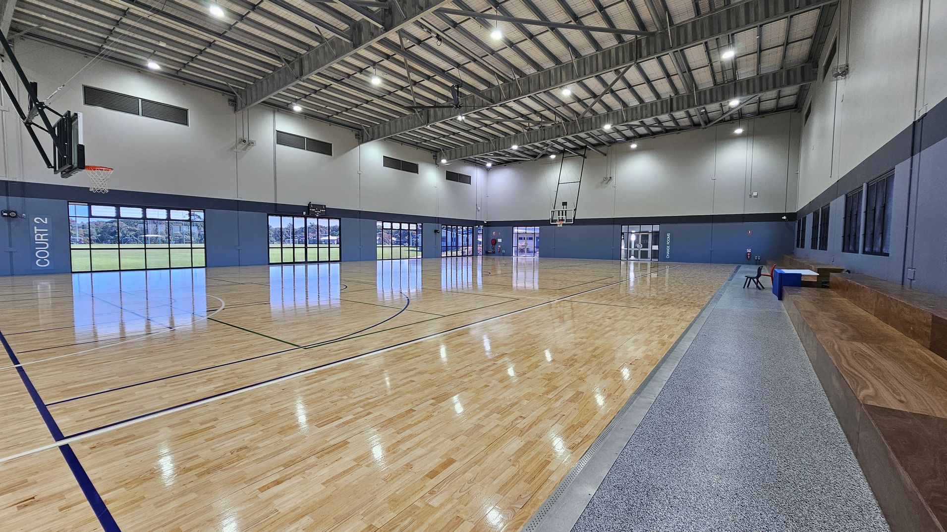 In a spacious sports hall, polished wooden flooring extends across the area, marked with blue and white lines for court play. On the left side, a basketball hoop is mounted above a grey wall, while "COURT 2" is clearly marked beneath large windows that provide views of the outside. To the right, glass doors lead to an entrance, and a raised wooden seating area runs along the wall, providing a place for spectators. The ceiling is high with exposed metal beams and bright overhead lights. In a spacious sports hall, polished wooden flooring extends across the area, marked with blue and white lines for court play. On the left side, a basketball hoop is mounted above a grey wall, while "COURT 2" is clearly marked beneath large windows that provide views of the outside. To the right, glass doors lead to an entrance, and a raised wooden seating area runs along the wall, providing a place for spectators. The ceiling is high with exposed metal beams and bright overhead lights.