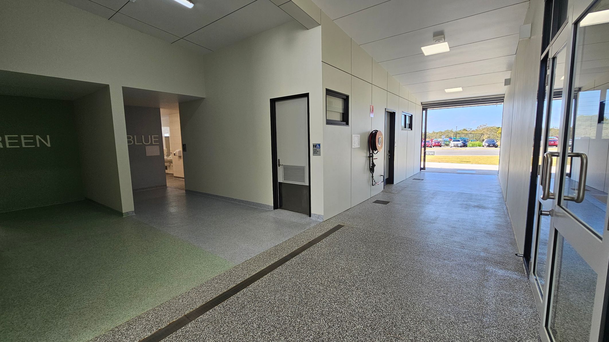 In a corridor, the floor features a textured combination of green and grey surfaces, smoothly transitioning towards a polished concrete section at the far end. On the left, a wall painted in a muted green displays the word "GREEN," with an adjoining door leading to another room. To the right, another wall marked with "BLUE" has a door beside it, which opens into a space with additional facilities. The ceiling is flat and white, fitted with square fluorescent lights. In a corridor, the floor features a textured combination of green and grey surfaces, smoothly transitioning towards a polished concrete section at the far end. On the left, a wall painted in a muted green displays the word "GREEN," with an adjoining door leading to another room. To the right, another wall marked with "BLUE" has a door beside it, which opens into a space with additional facilities. The ceiling is flat and white, fitted with square fluorescent lights.