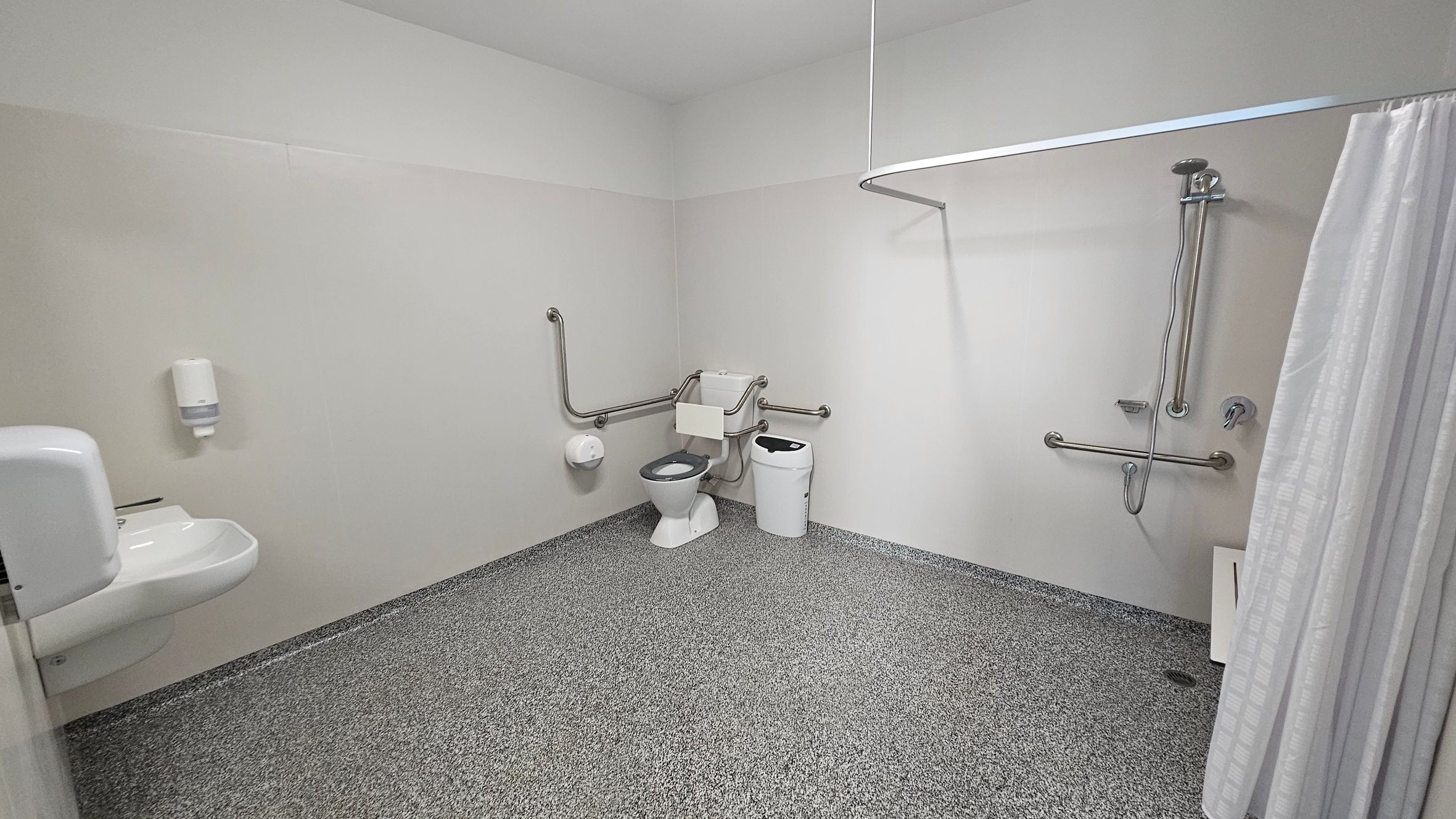 The room features a textured grey floor that extends throughout the space, providing a non-slip surface. On the left, a white sink is mounted on the wall with a soap dispenser above it. Adjacent to this is a toilet, equipped with grab bars for accessibility. On the right side, a shower area is enclosed by a curtain, with additional grab bars mounted on the wall next to the shower head. A small, rectangular waste bin sits beside the toilet, and the ceiling is flat and finished in white. The room features a textured grey floor that extends throughout the space, providing a non-slip surface. On the left, a white sink is mounted on the wall with a soap dispenser above it. Adjacent to this is a toilet, equipped with grab bars for accessibility. On the right side, a shower area is enclosed by a curtain, with additional grab bars mounted on the wall next to the shower head. A small, rectangular waste bin sits beside the toilet, and the ceiling is flat and finished in white.