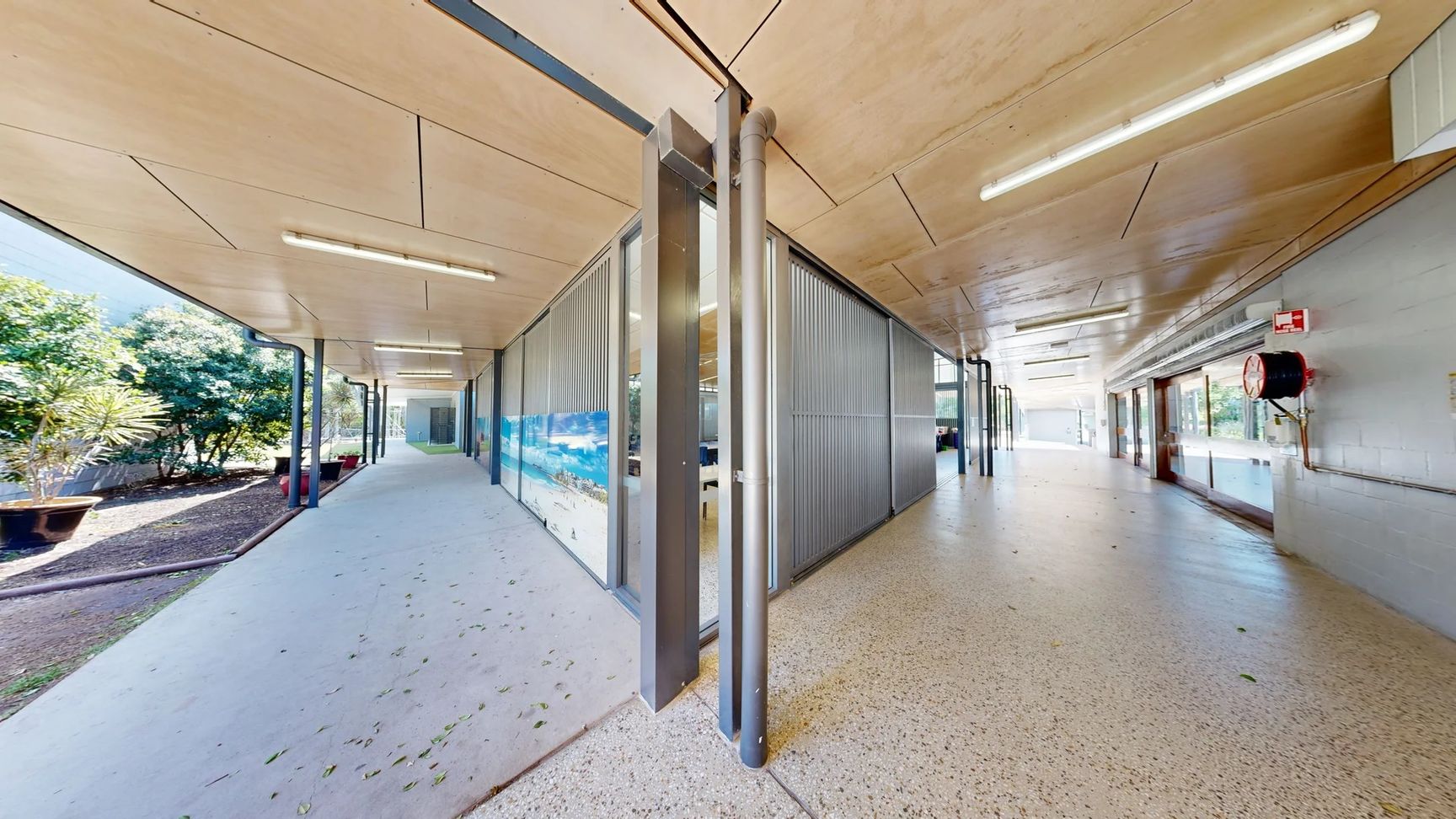 On the left, a sloped pathway leads towards a smooth concrete surface bordered by a low wall. There’s a white door with a metal handle and a small sign next to it. Mounted above, there are multiple fire alarms and a wall sign with safety information. On the right, a series of glass panels reveal a corridor with additional doors further down. The floor is a light grey concrete, and the ceiling is flat, featuring wooden panels with fluorescent lighting fixtures spaced evenly. On the left, a sloped pathway leads towards a smooth concrete surface bordered by a low wall. There’s a white door with a metal handle and a small sign next to it. Mounted above, there are multiple fire alarms and a wall sign with safety information. On the right, a series of glass panels reveal a corridor with additional doors further down. The floor is a light grey concrete, and the ceiling is flat, featuring wooden panels with fluorescent lighting fixtures spaced evenly.