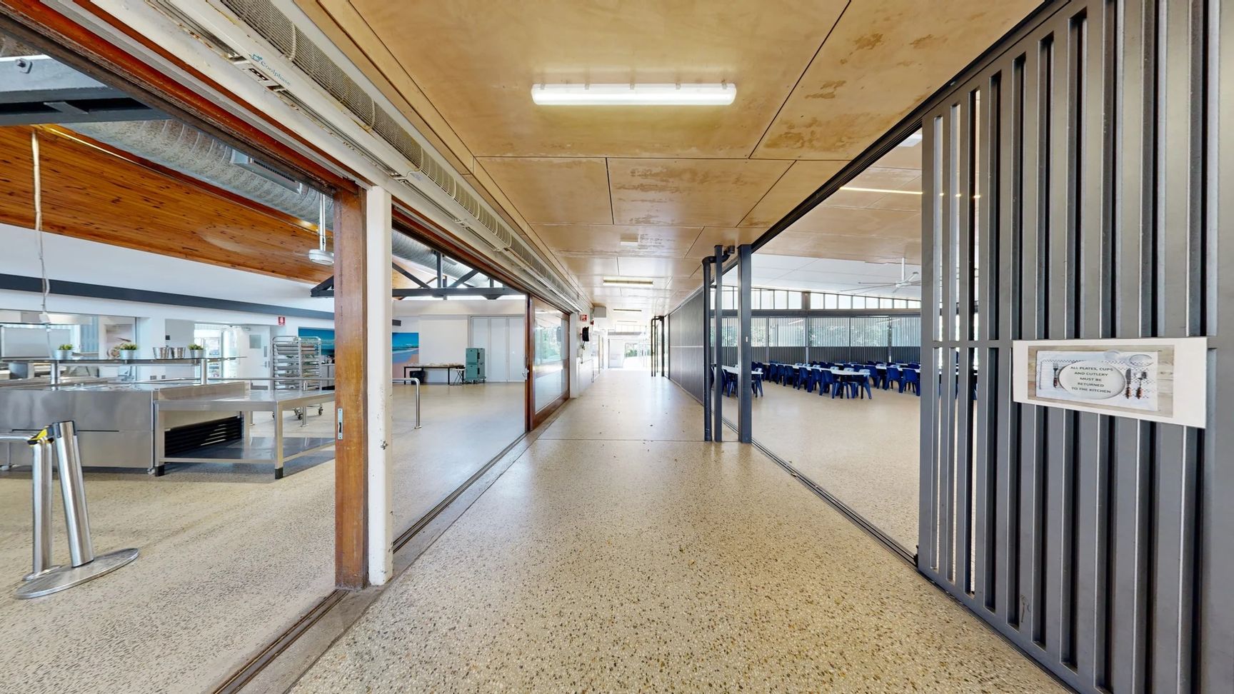 On the left, there is a wide entrance leading into a kitchen area with stainless steel equipment and shelving visible, along with a light-coloured wall. A smooth, polished floor extends throughout. On the right, a grey partition separates the corridor from a dining area filled with rows of blue seats around tables. The ceiling features a combination of flat panels and exposed beams, with overhead lights illuminating the space. On the left, there is a wide entrance leading into a kitchen area with stainless steel equipment and shelving visible, along with a light-coloured wall. A smooth, polished floor extends throughout. On the right, a grey partition separates the corridor from a dining area filled with rows of blue seats around tables. The ceiling features a combination of flat panels and exposed beams, with overhead lights illuminating the space.