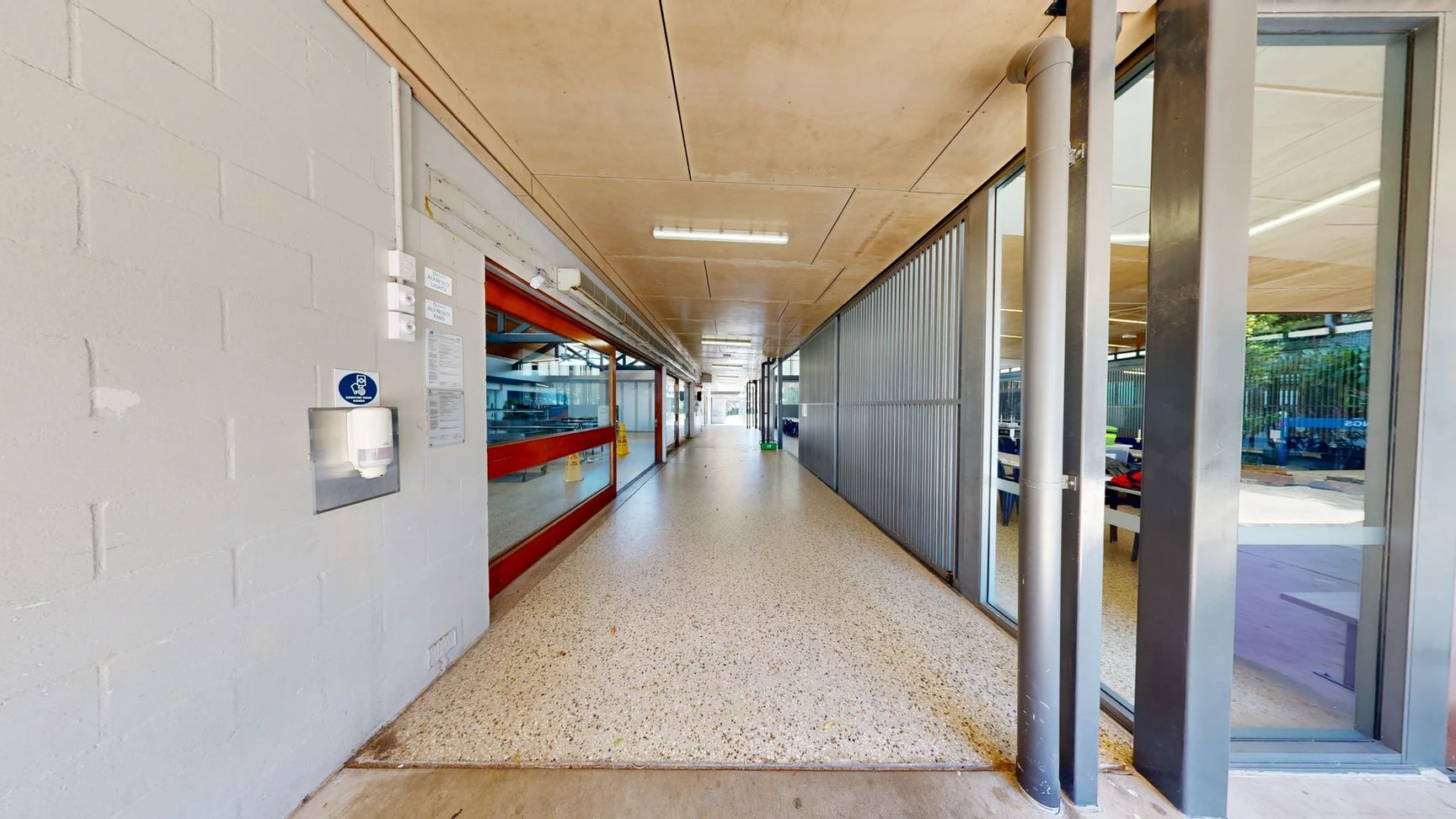 On the left, a light grey concrete wall features a paper towel dispenser and several mounted notices. A red-framed window offers a view into a room with equipment. The floor is a smooth, speckled surface that transitions to wood near the edge. On the right, large glass panels are set in a steel frame, revealing a corridor with blue tables and chairs in the background. The ceiling is a flat concrete design with strip lighting running along its length. On the left, a light grey concrete wall features a paper towel dispenser and several mounted notices. A red-framed window offers a view into a room with equipment. The floor is a smooth, speckled surface that transitions to wood near the edge. On the right, large glass panels are set in a steel frame, revealing a corridor with blue tables and chairs in the background. The ceiling is a flat concrete design with strip lighting running along its length.
