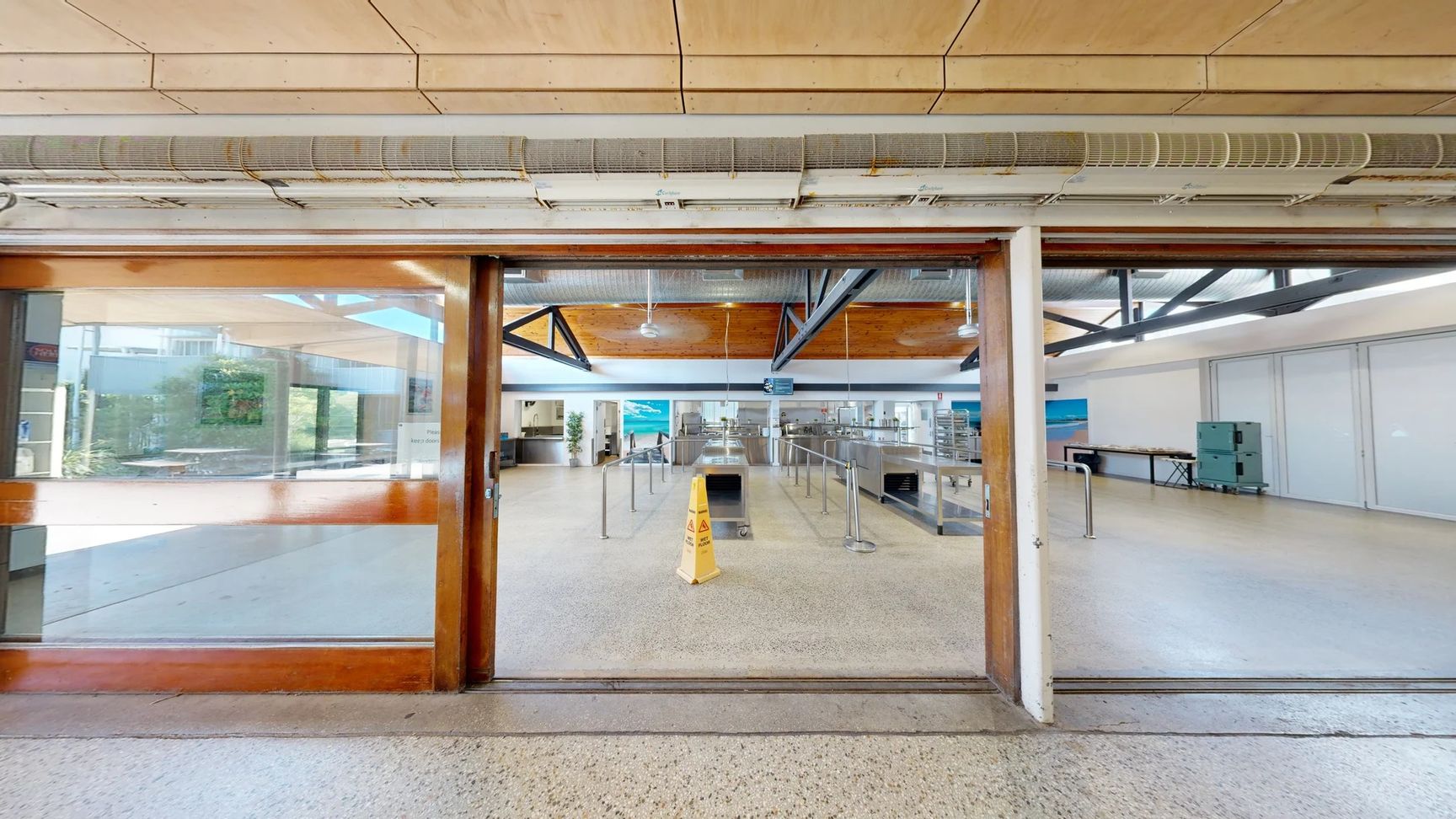 On the left, there’s a large glass door with wooden frames leading to an outdoor area visible through the glass. A small sign is attached to the wall nearby. To the right, a hallway opens into a kitchen area with stainless steel counters and equipment. The floor is smooth, speckled concrete, and the ceiling features wooden panels with exposed beams and overhead lights. In the centre, a caution sign is placed on the floor near a line of barriers guiding visitors towards the service area. On the left, there’s a large glass door with wooden frames leading to an outdoor area visible through the glass. A small sign is attached to the wall nearby. To the right, a hallway opens into a kitchen area with stainless steel counters and equipment. The floor is smooth, speckled concrete, and the ceiling features wooden panels with exposed beams and overhead lights. In the centre, a caution sign is placed on the floor near a line of barriers guiding visitors towards the service area.