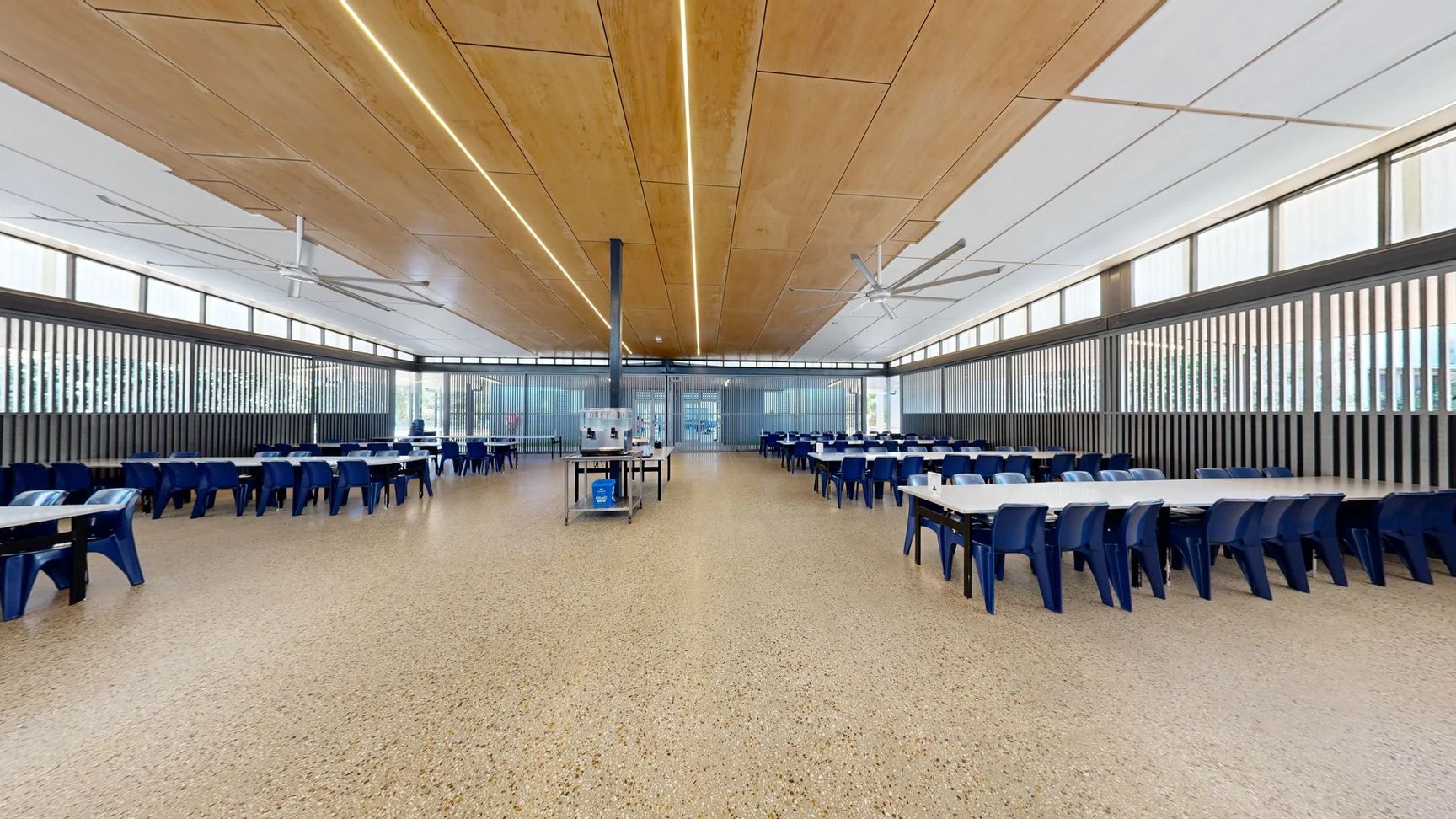 On the left, a row of blue tables and chairs extends along the wall, with additional seating visible further down. On the right, more tables with blue chairs create a symmetrical layout, with narrow vertical windows above. In the centre, a serving cart stands on a polished, speckled concrete floor. The ceiling features wooden panels with integrated lighting and large ceiling fans. The space is well-lit, and the layout is open, with clear pathways between tables leading to a background area. On the left, a row of blue tables and chairs extends along the wall, with additional seating visible further down. On the right, more tables with blue chairs create a symmetrical layout, with narrow vertical windows above. In the centre, a serving cart stands on a polished, speckled concrete floor. The ceiling features wooden panels with integrated lighting and large ceiling fans. The space is well-lit, and the layout is open, with clear pathways between tables leading to a background area.