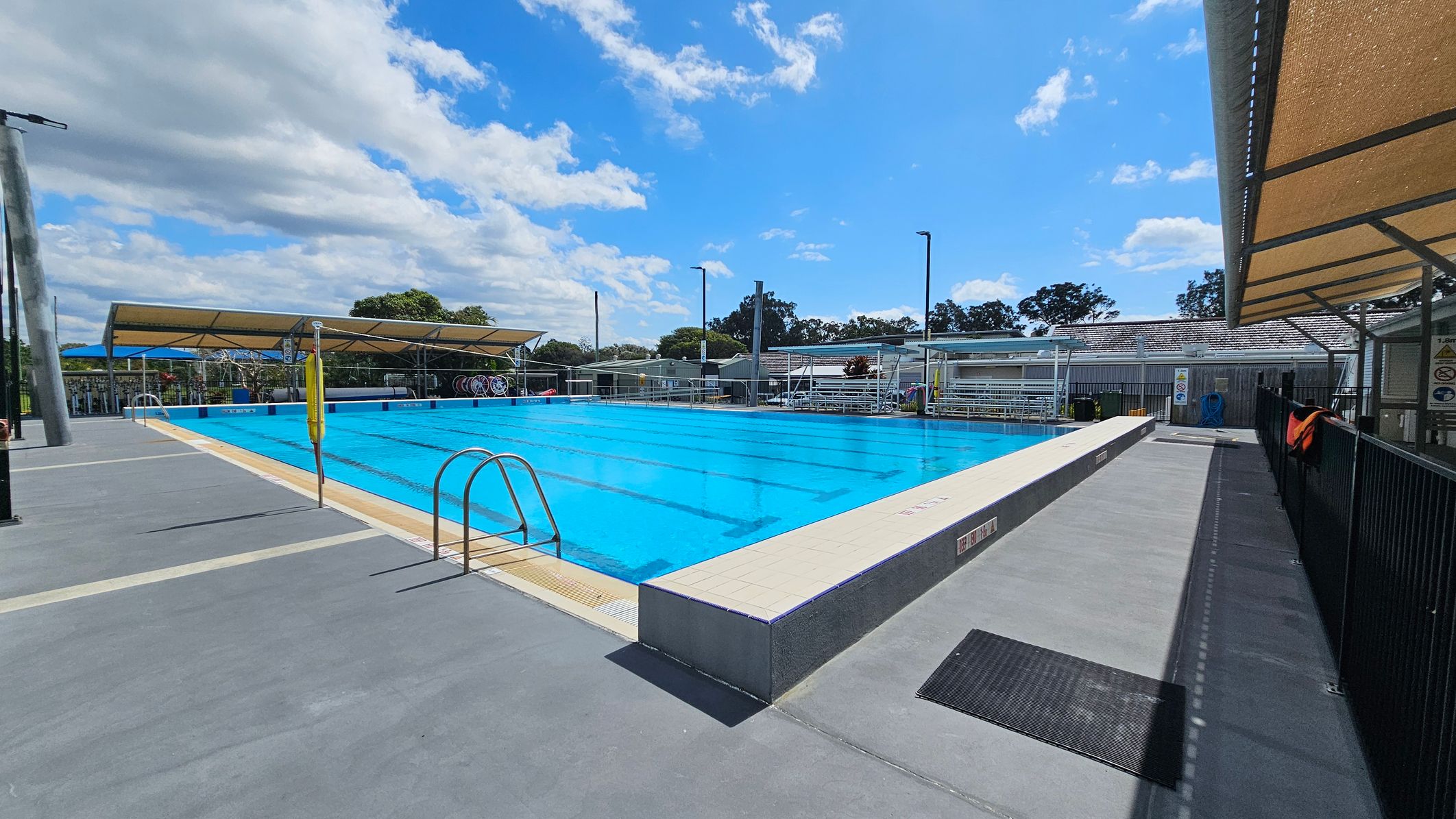 On the left, a row of large green shrubs adds a natural element beside a white building with vents and a smooth, light-coloured wall. To the right, a shaded area features a metal frame with a canopy, providing coverage over a walkway. The pool is visible in the middle, with clear blue water and a light-coloured edge. The ground is a clean, pale surface, and there are metal fences marking the pool area. On the left, a row of large green shrubs adds a natural element beside a white building with vents and a smooth, light-coloured wall. To the right, a shaded area features a metal frame with a canopy, providing coverage over a walkway. The pool is visible in the middle, with clear blue water and a light-coloured edge. The ground is a clean, pale surface, and there are metal fences marking the pool area.