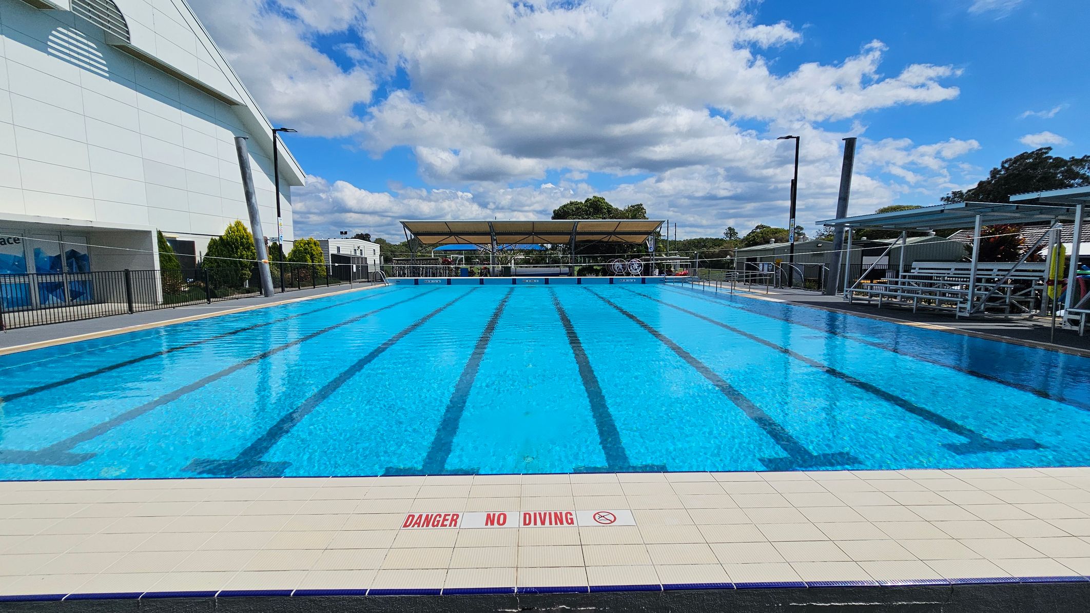 An outdoor swimming pool with clear blue water and marked lanes. The pool is surrounded by a tiled deck with a "Danger No Diving" warning. On the left, there's a large building with a slanted roof and some greenery. On the right, there are metal bleachers under a canopy. In the background, trees and a covered area with equipment are visible. The sky is partly cloudy. An outdoor swimming pool with clear blue water and marked lanes. The pool is surrounded by a tiled deck with a "Danger No Diving" warning. On the left, there's a large building with a slanted roof and some greenery. On the right, there are metal bleachers under a canopy. In the background, trees and a covered area with equipment are visible. The sky is partly cloudy.