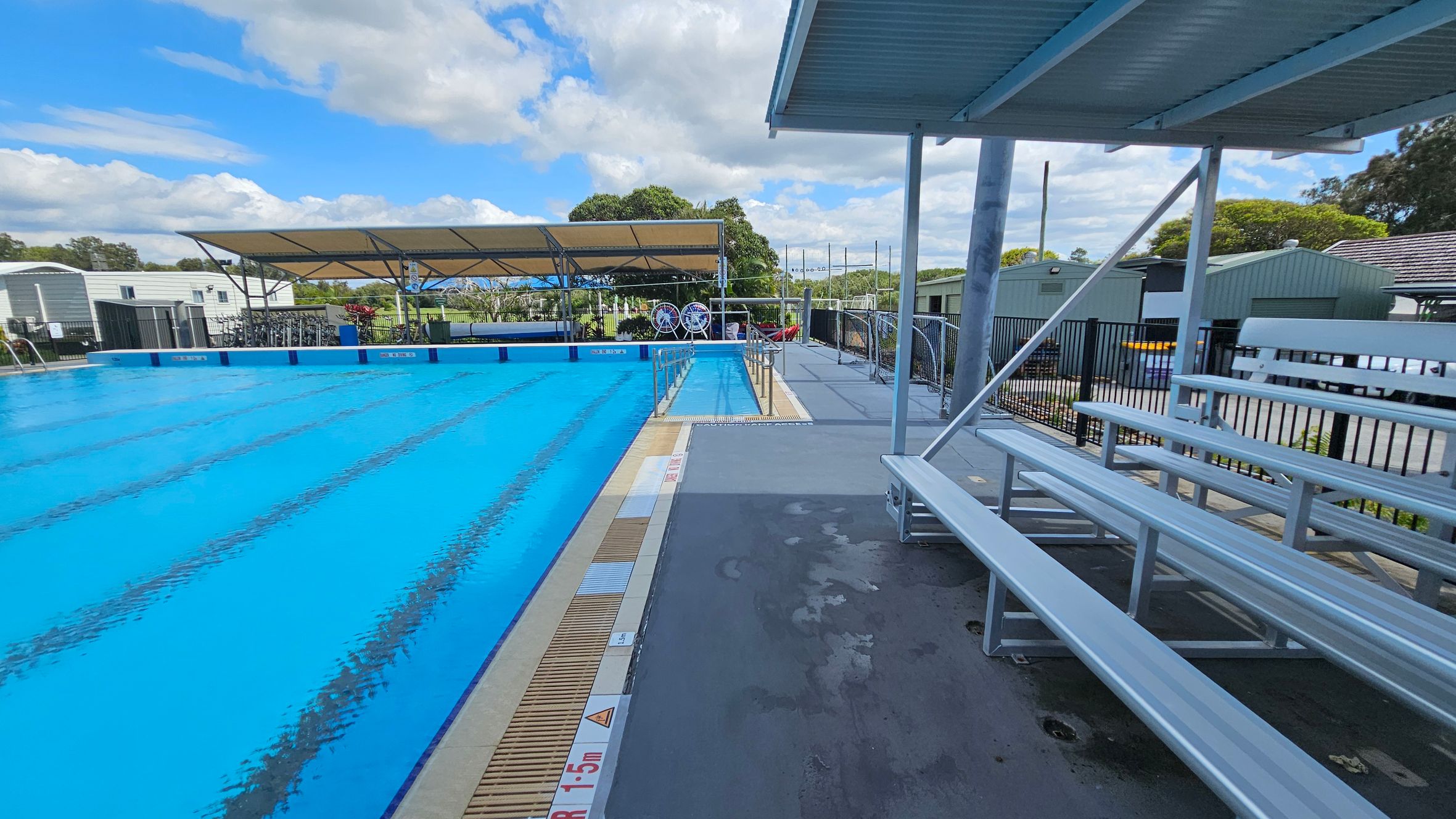 On the left, there's a large swimming pool with clear blue water, bordered by a white building with a slanted roof. A yellow flotation device hangs on a pole by the poolside. On the right, there are metal bleachers under a corrugated roof, with a bin filled with colourful pool noodles. In the background, there are some plants and a fence, with a few structures visible beyond the pool area. On the left, there's a large swimming pool with clear blue water, bordered by a white building with a slanted roof. A yellow flotation device hangs on a pole by the poolside. On the right, there are metal bleachers under a corrugated roof, with a bin filled with colourful pool noodles. In the background, there are some plants and a fence, with a few structures visible beyond the pool area.