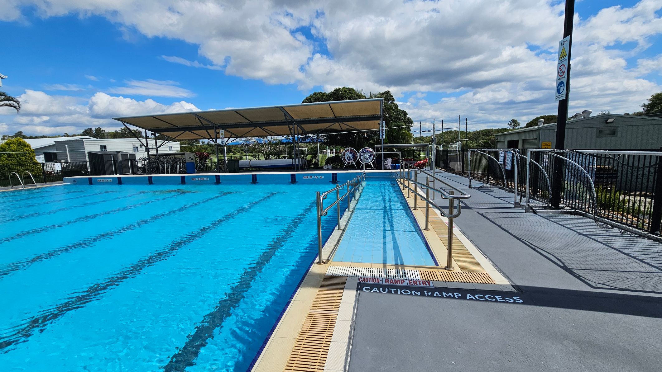 An outdoor swimming pool with clear blue water and lane markings. On the right, there's a ramp with metal railings leading into the pool, marked with "Caution Ramp Access." A shaded area with a canopy is in the background, along with some buildings and trees. The pool deck is grey with a black fence running along the perimeter. Signs are visible on a pole near the fence. An outdoor swimming pool with clear blue water and lane markings. On the right, there's a ramp with metal railings leading into the pool, marked with "Caution Ramp Access." A shaded area with a canopy is in the background, along with some buildings and trees. The pool deck is grey with a black fence running along the perimeter. Signs are visible on a pole near the fence.